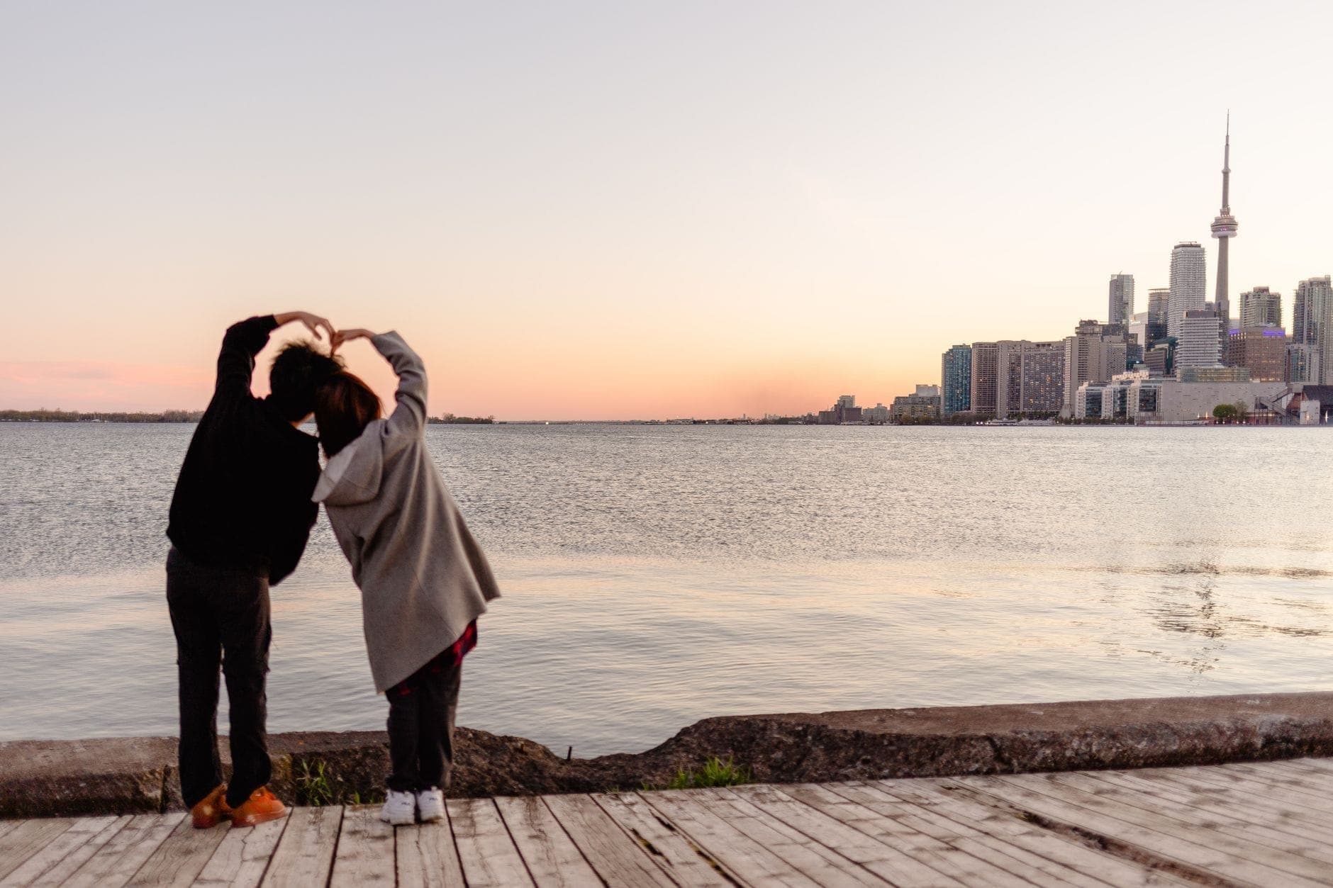 Couple making a heart shape with their arms while standing by Lake Ontario with the Toronto skyline at sunset