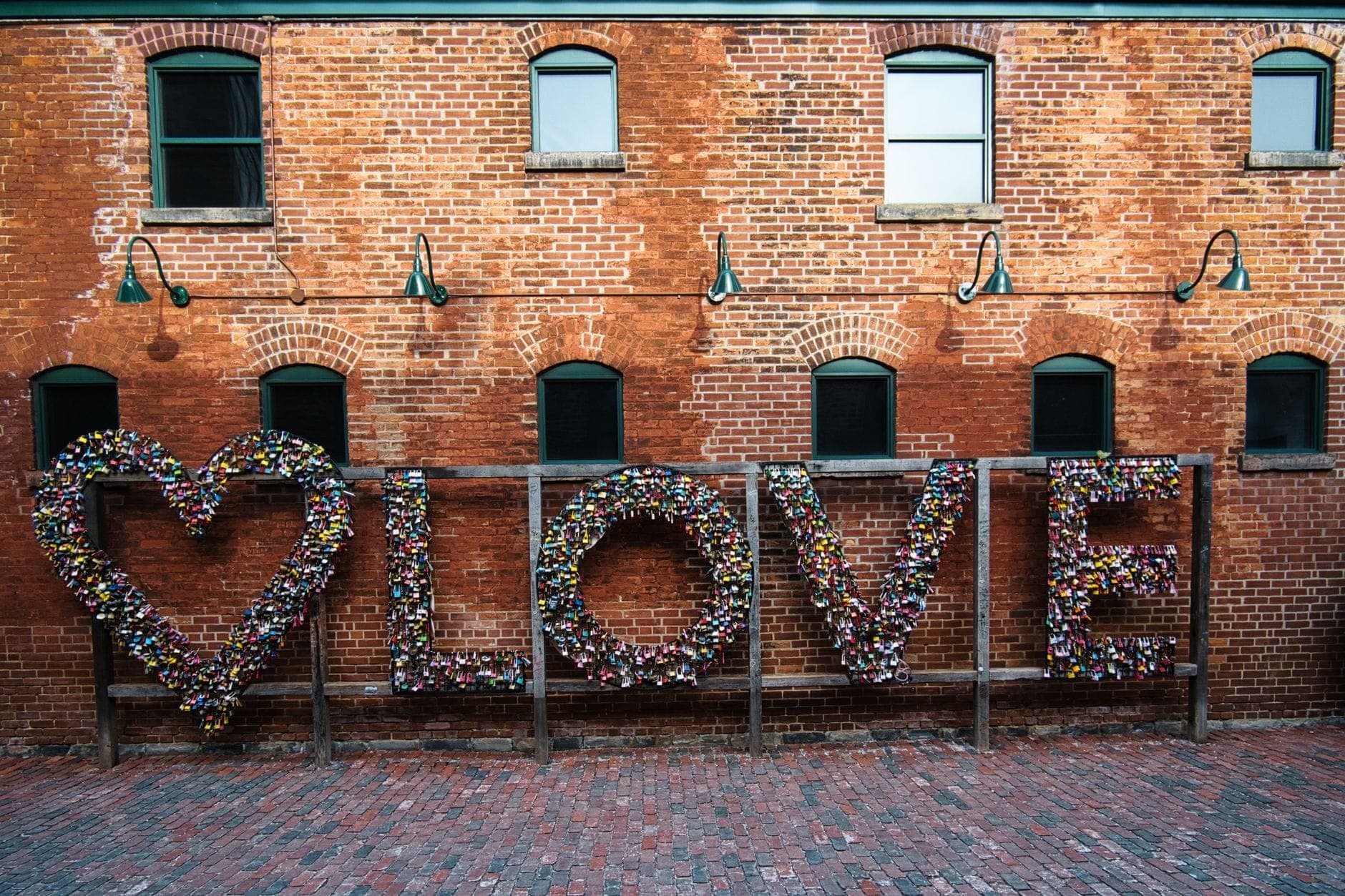 Large LOVE sign covered with locks against a red brick wall and cobblestone lane in Toronto’s Distillery District.