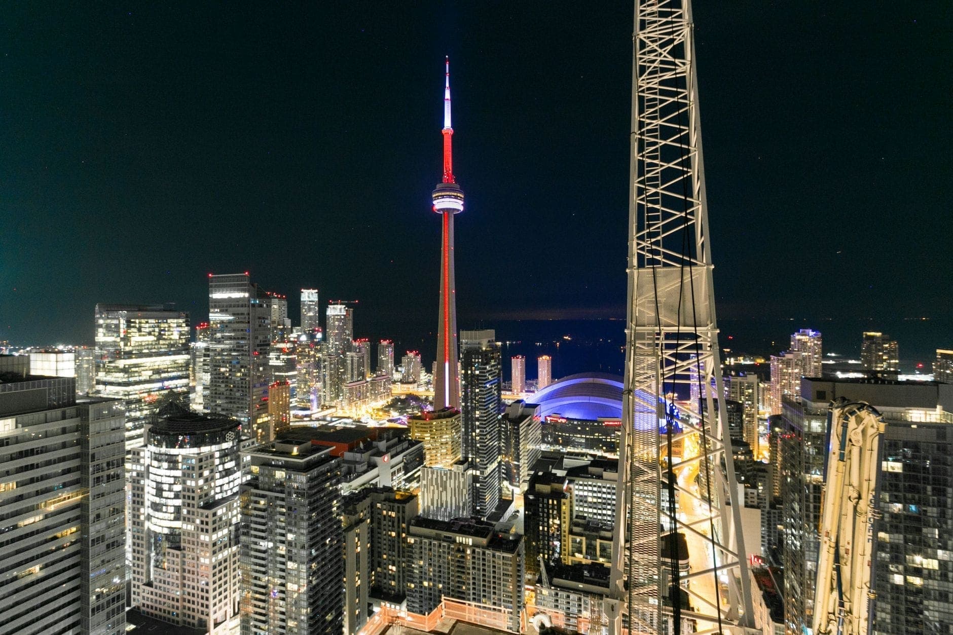Nighttime view of Toronto skyline with the CN Tower prominently lit, surrounded by tall buildings and city lights, seen from a high vantage point.