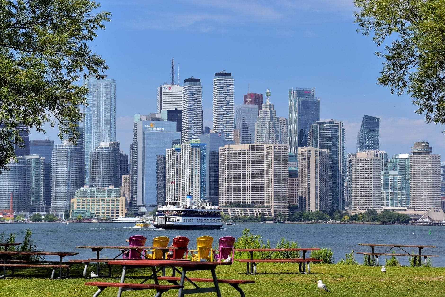 Picnic benches and colorful Muskoka chairs overlook Toronto’s skyline, seen across the water from the Toronto Islands on a sunny day.