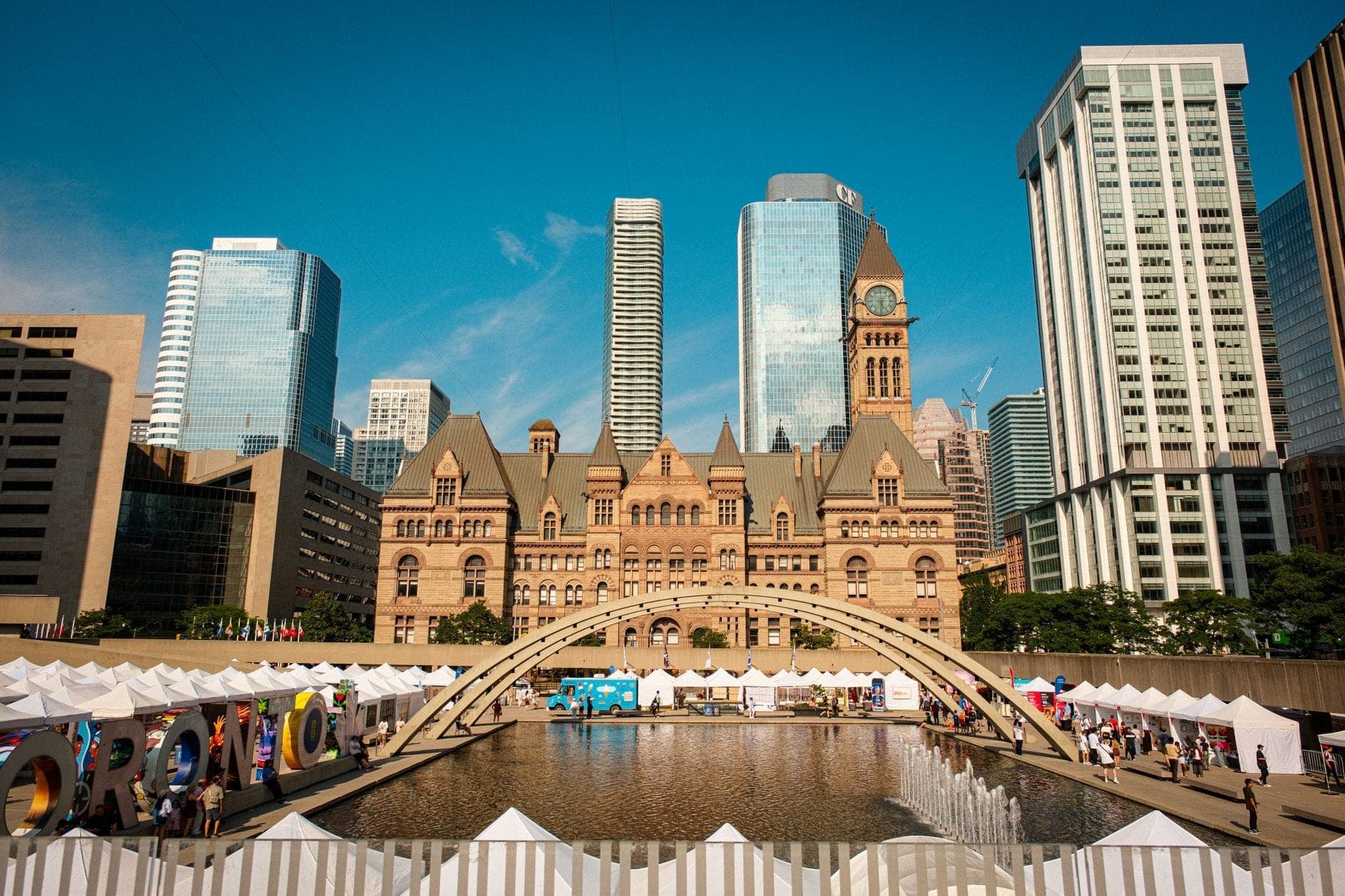 Toronto’s Old City Hall and modern skyscrapers overlook summer festival tents and a reflecting pool at Nathan Phillips Square.