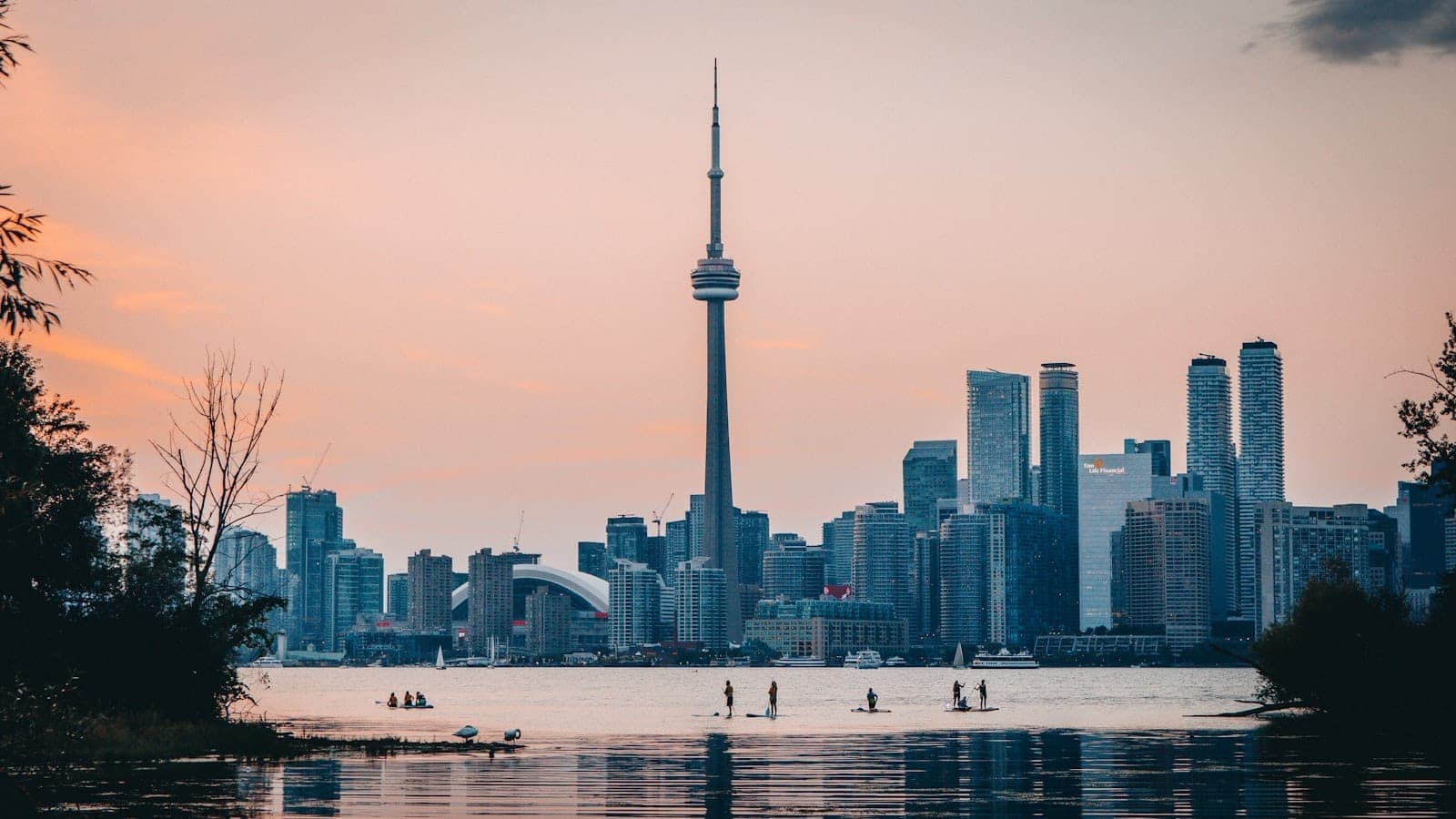 Lo skyline di Toronto al tramonto visto dalle Isole di Toronto, con l'acqua calma, gli alberi che incorniciano il panorama e alcune persone che fanno paddleboard in primo piano.