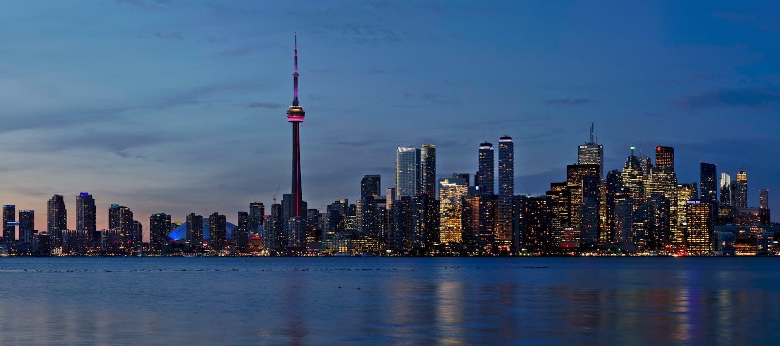 Toronto skyline at dusk with the CN Tower and city lights reflecting on Lake Ontario under a blue evening sky.