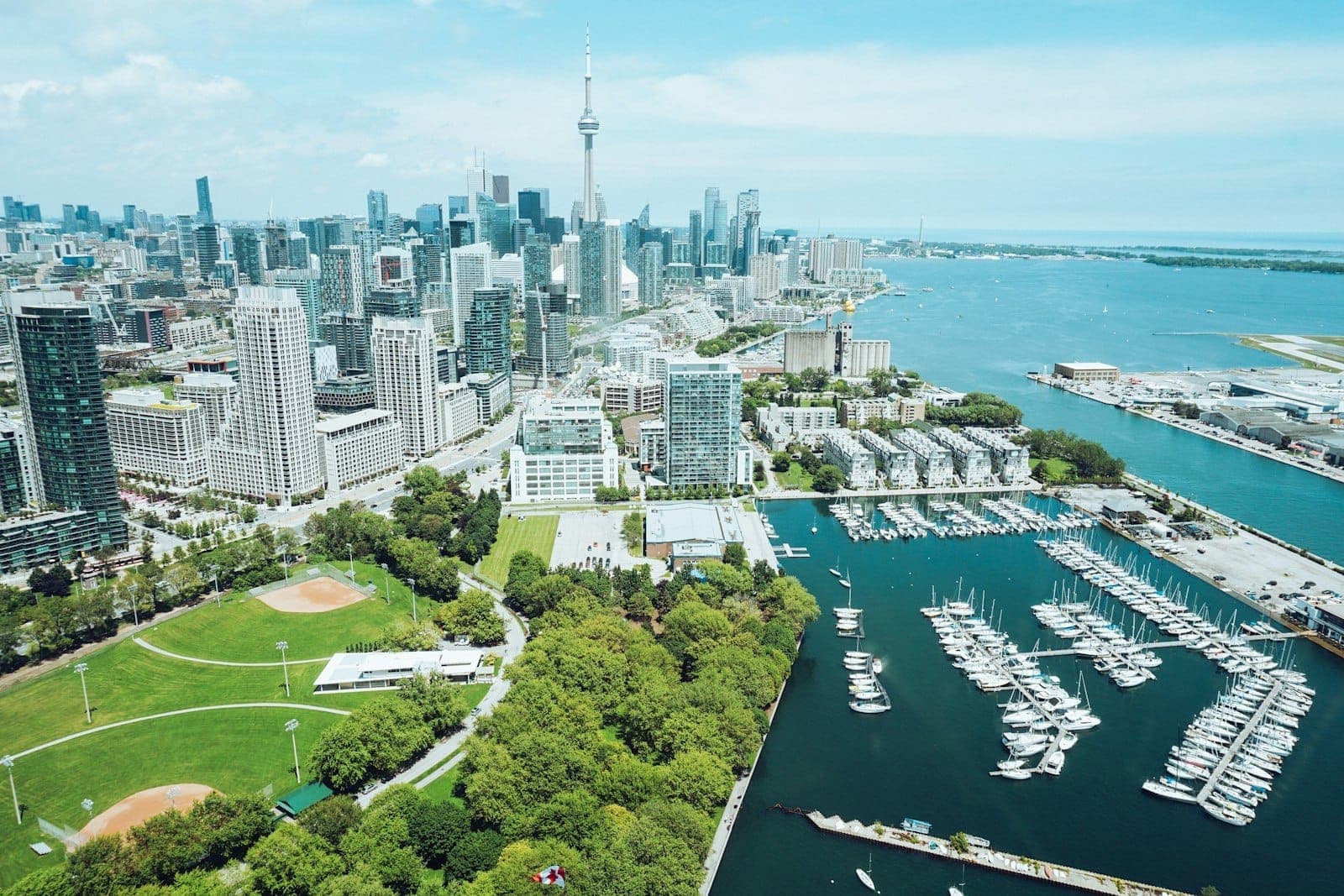 Vista aerea del Toronto Waterfront con lo skyline del centro città, la CN Tower, il porto con barche ormeggiate, campi sportivi e rigogliose aree verdi sotto un cielo sereno.