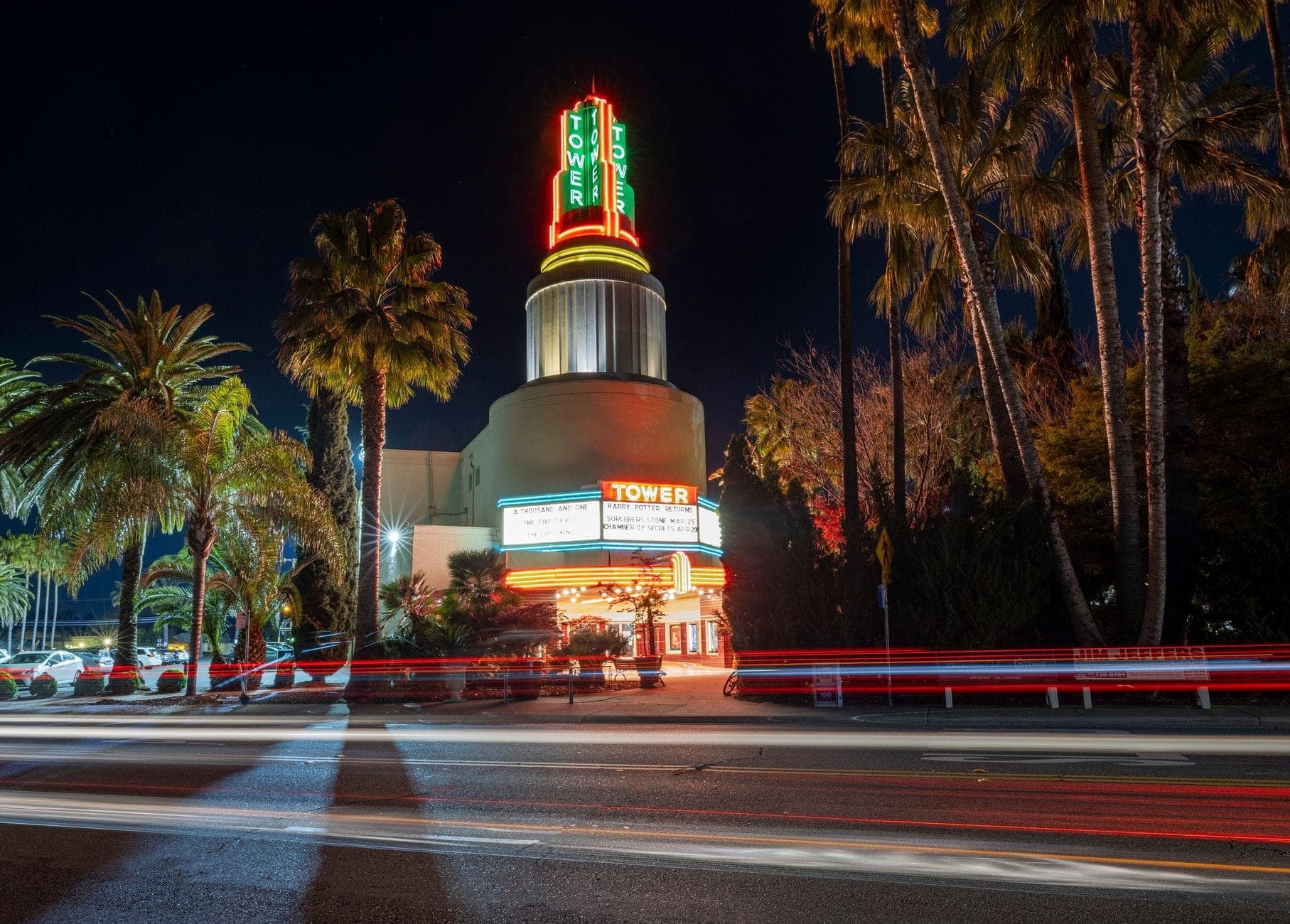 Night view of an art deco cinema with a neon-lit marquee tower reading TOWER, surrounded by palm trees and light trails on a busy street.