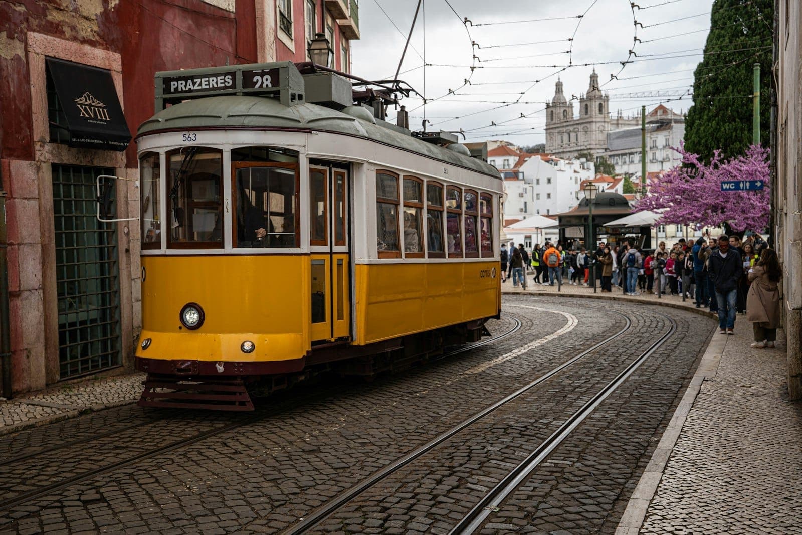 O clássico Elétrico 28E amarelo faz uma curva em uma rua de paralelepípedos em Lisboa, com prédios antigos, flores da primavera e turistas ao fundo.