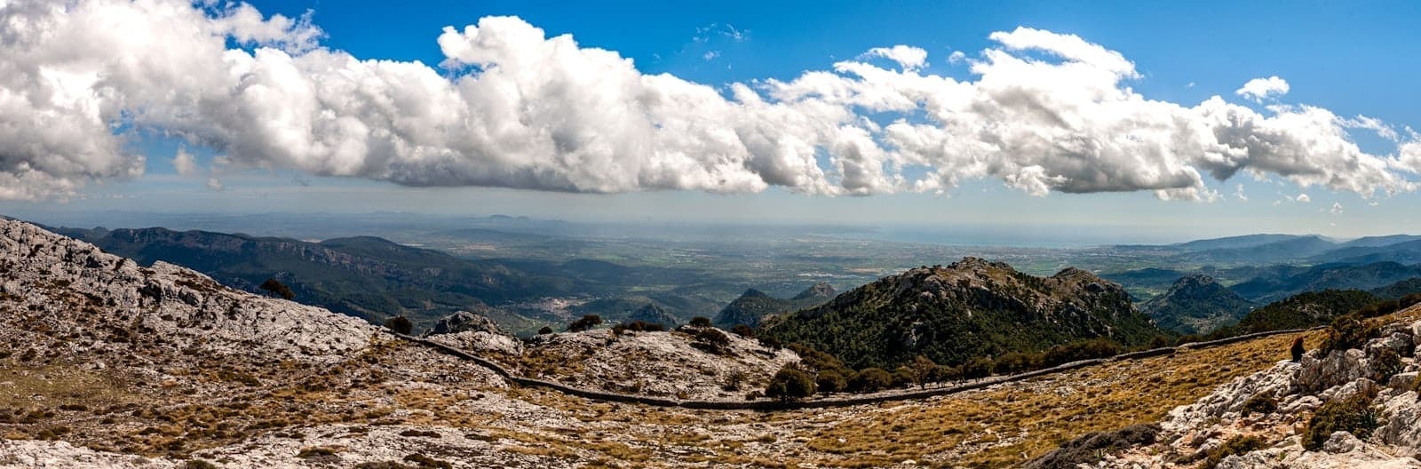 Panoramablick auf die Serra de Tramuntana mit markanten Kalksteingipfeln, einer kurvenreichen Straße und einem weiten Tal unter strahlend blauem Himmel mit vereinzelten Wolken.