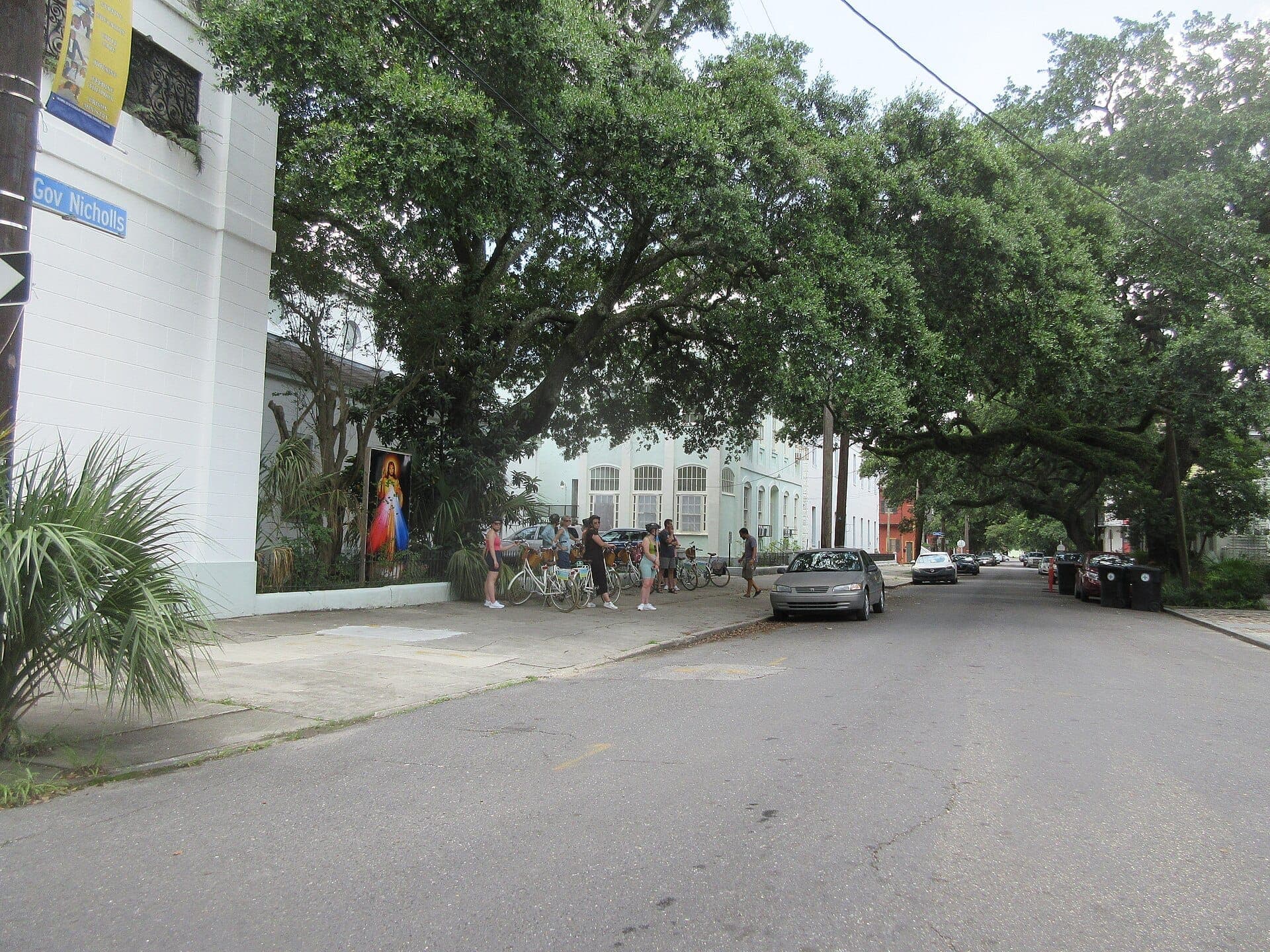 Street view in Tremé featuring large oak trees, pastel buildings, parked cars, and a few people with bicycles on the sidewalk.