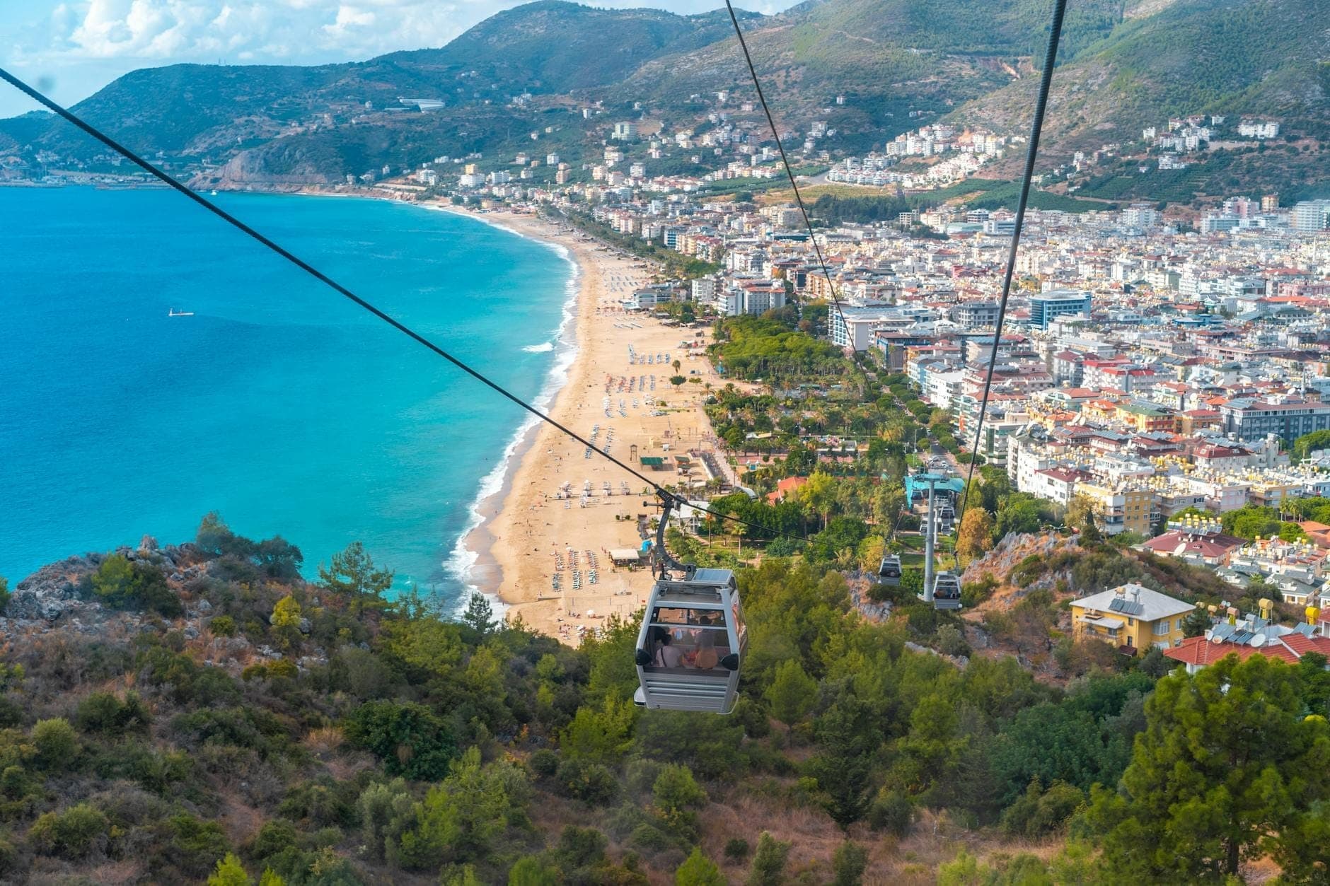 Eine Gondel schwebt über saftig grüne Hügel mit einem weiten Blick auf Antalyas Konyaaltı-Strand, die Stadtsilhouette und die fernen Taurusberge unter blauem Himmel.