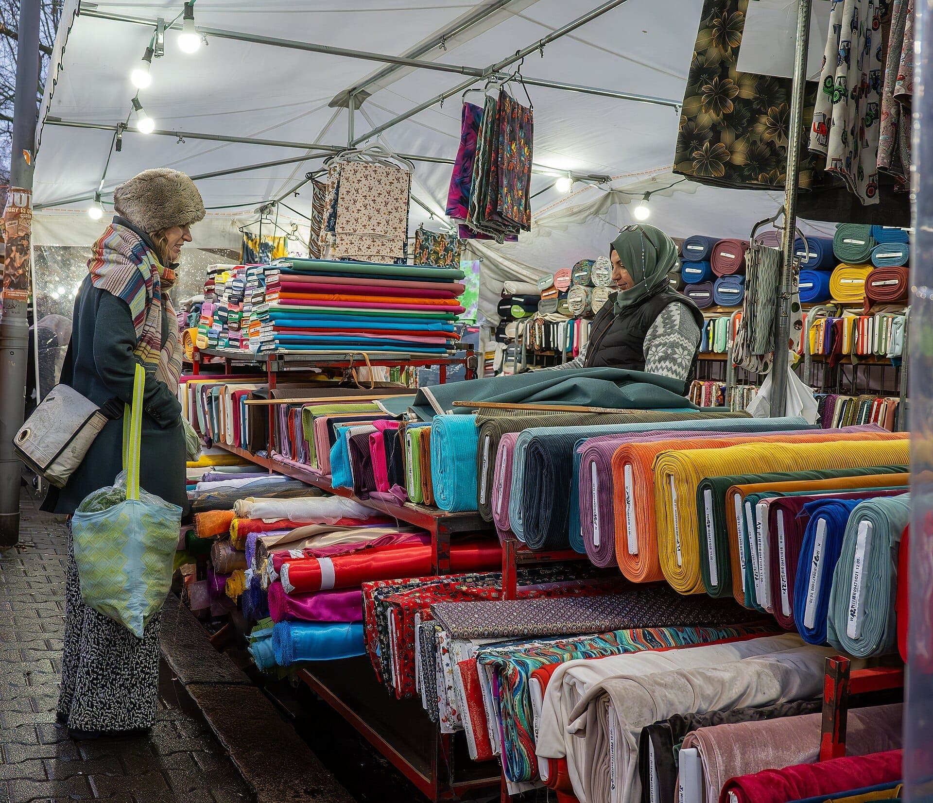 ม้วนผ้าหลากสีสันที่วางแสดงอยู่ในแผงขายที่คึกคักของ Turkish Market Maybachufer โดยมีผู้หญิงสองคนกำลังเลือกดูสินค้าใต้หลังคาสีขาว