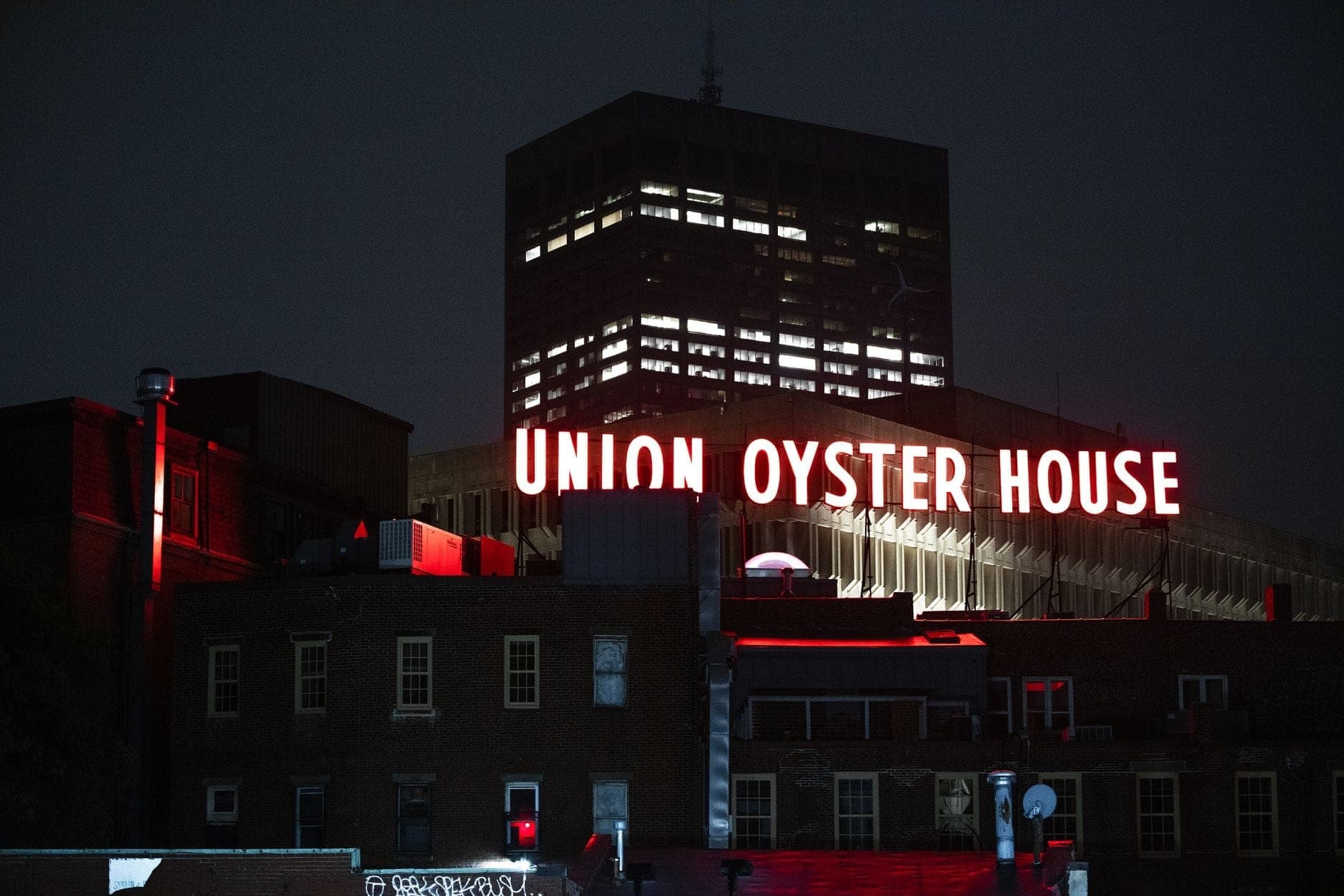 Vue nocturne de l'enseigne au néon de l'Union Oyster House brillant au-dessus des bâtiments en briques historiques de Boston, avec une tour de bureaux moderne en arrière-plan.