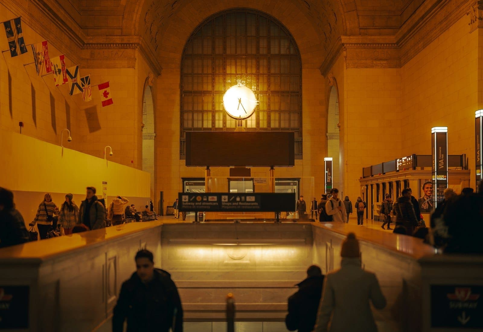 Interno della Great Hall della Union Station di Toronto al crepuscolo, con persone in cammino, un orologio illuminato, alte finestre ad arco, pareti in calcare e bandiere che sventolano in alto.