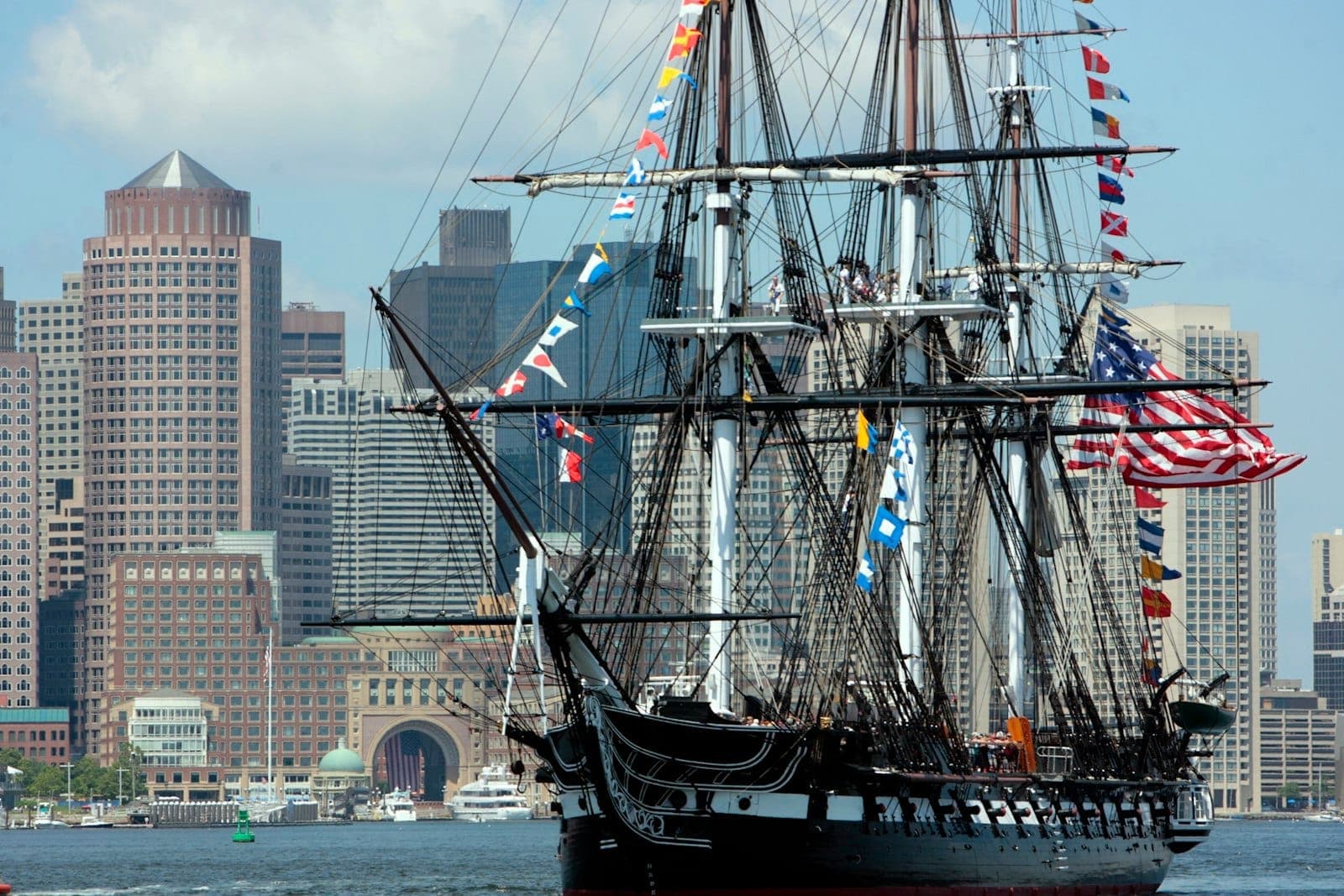 USS Constitution, orné de pavillons de signalisation colorés et d'un grand drapeau américain, amarré dans le port de Boston avec la ligne d'horizon de la ville en arrière-plan.