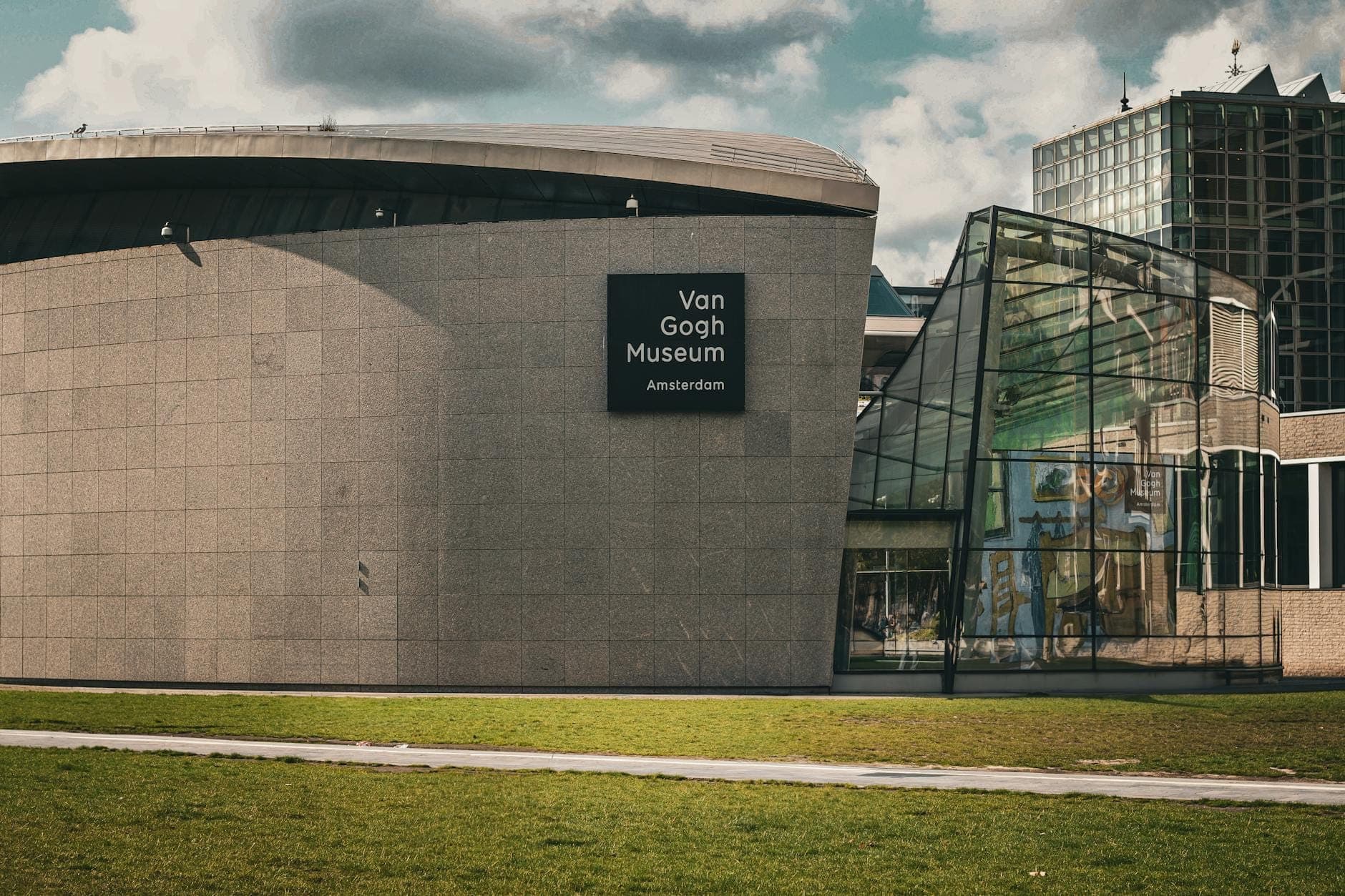 Die Außenansicht des Van Gogh Museums in Amsterdam mit seiner markanten modernen Architektur, dem Eingangsbereich und einer grünen Rasenfläche unter leicht bewölktem Himmel.