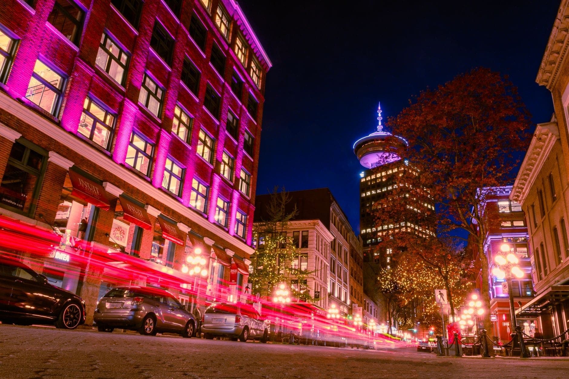 ถนนคึกคักในย่าน Gastown ช่วงกลางคืน แสงไฟอาคารชมพู เหลือง โคมไฟถนนสว่าง และ Harbour Centre เปิดไฟโดดเด่นอยู่ไกล ๆ