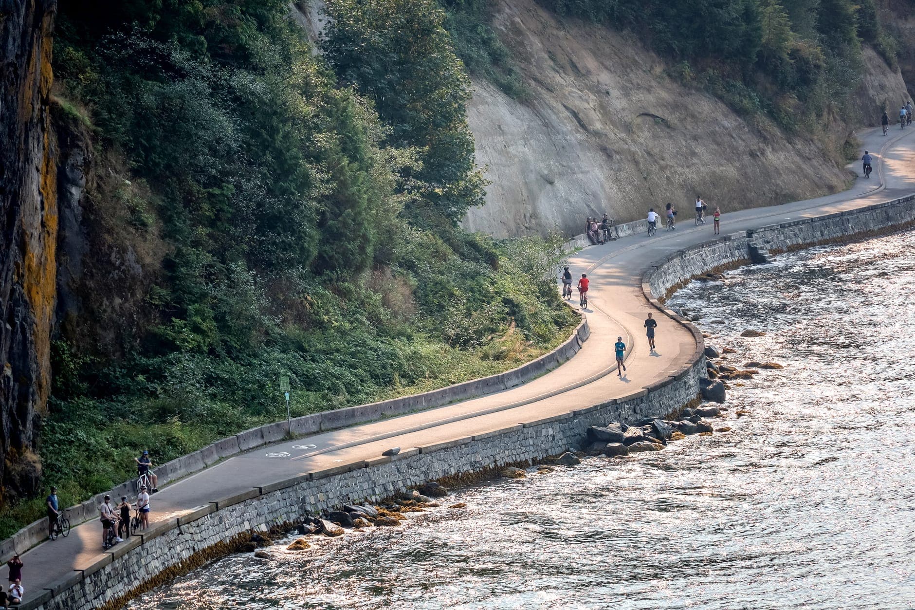 ทางโค้งริมทะเลของ Vancouver Seawall มีผู้คนเดินและปั่นจักรยานข้างมหาสมุทร ติดหน้าผาและต้นไม้เขียวขจีในวันที่แดดจัด