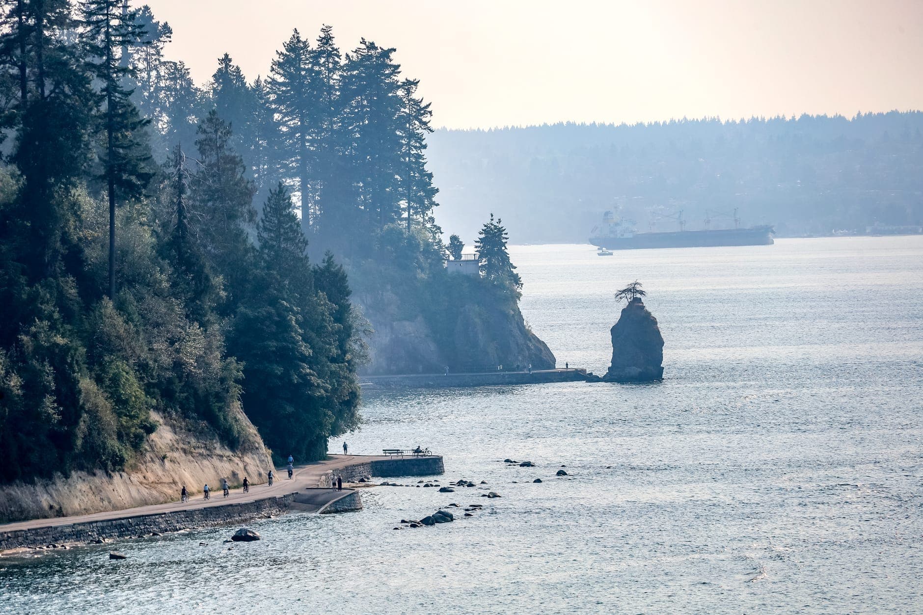 ทางเดิน Seawall ของ Stanley Park ในเมือง Vancouver พร้อมต้นไม้และมหาสมุทร เกาะหินเล็กๆ และเรือในระยะไกลในวันที่แสงสดใส