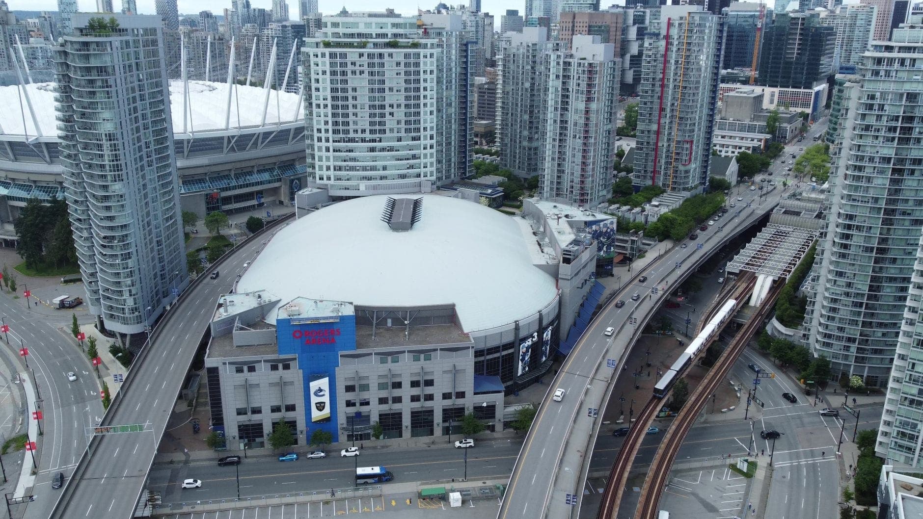 มุมมองทางอากาศของย่านดาวน์ทาวน์แวนคูเวอร์ พร้อมรางรถไฟ SkyTrain ขบวนรถไฟ Rogers Arena และสนามกีฬา BC Place ที่ล้อมรอบด้วยอาคารสูง