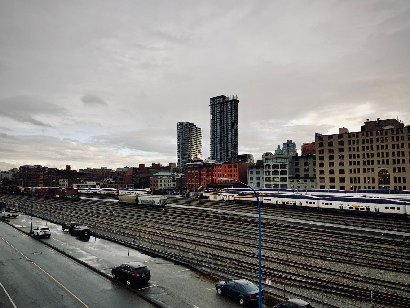 ลานรถไฟพร้อมรถไฟโดยสารและเส้นขอบฟ้าของเมือง น่าจะเป็น Vancouver ภายใต้ท้องฟ้าครึ้ม