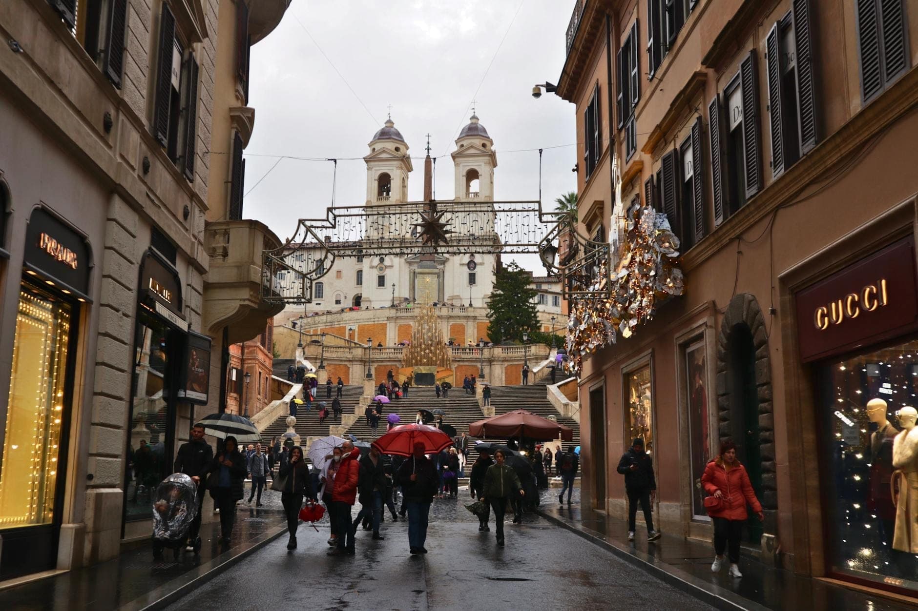Mga taong naglalakad sa Via dei Condotti sa makulimlim na araw, napapalibutan ng mga mamahaling tindahan tulad ng Gucci at mga palamuting ilaw, kasama ang Spanish Steps sa likuran.