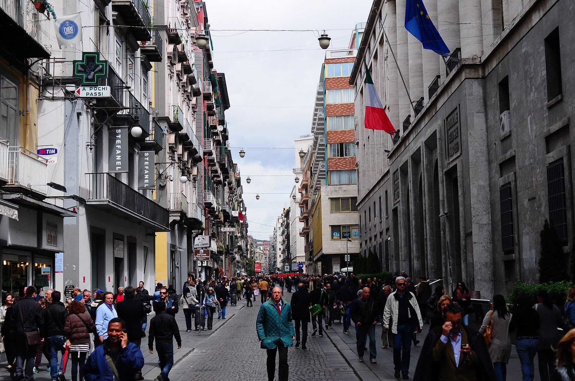 Multidão de pessoas caminha pela Via Toledo em Nápoles, ladeada por prédios históricos, lojas e bandeiras italianas numa animada rua de pedestres.