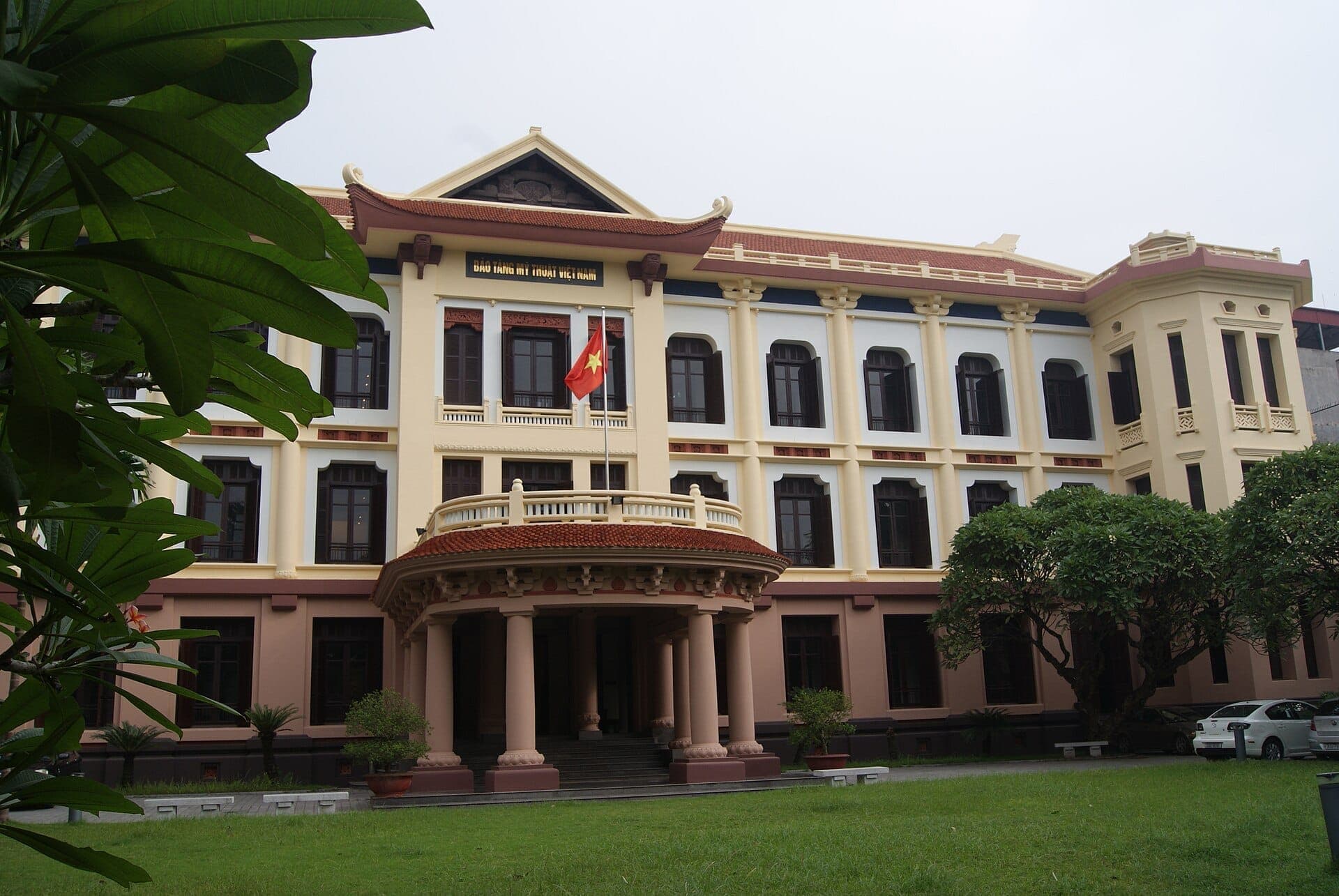Widok z przodu Vietnam Fine Arts Museum w Hanoi, prezentujący francuską architekturę kolonialną, flagę wietnamską i zielony ogród w pochmurny dzień.