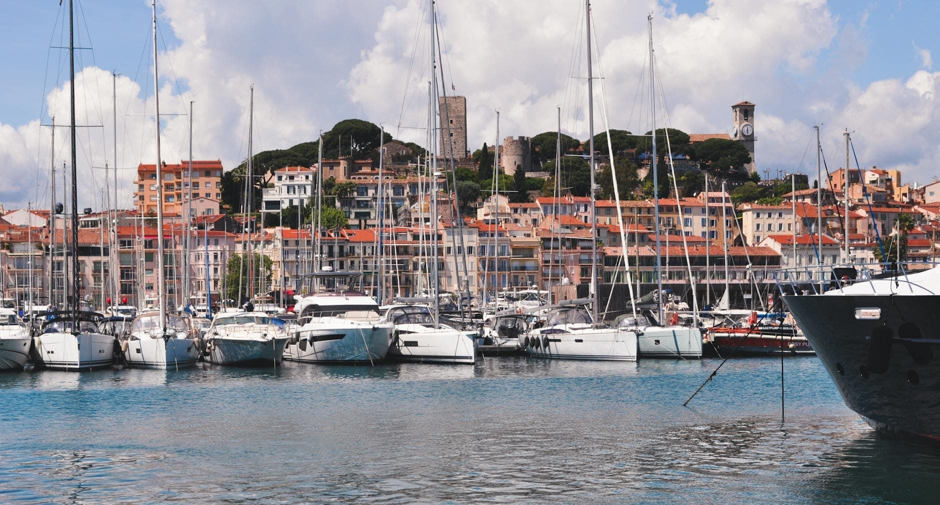 Панорамный вид на Vieux Port de Cannes: яхты и лодки у причала, средневековая башня Ле-Сюке и разноцветные дома старого города под ярким небом.