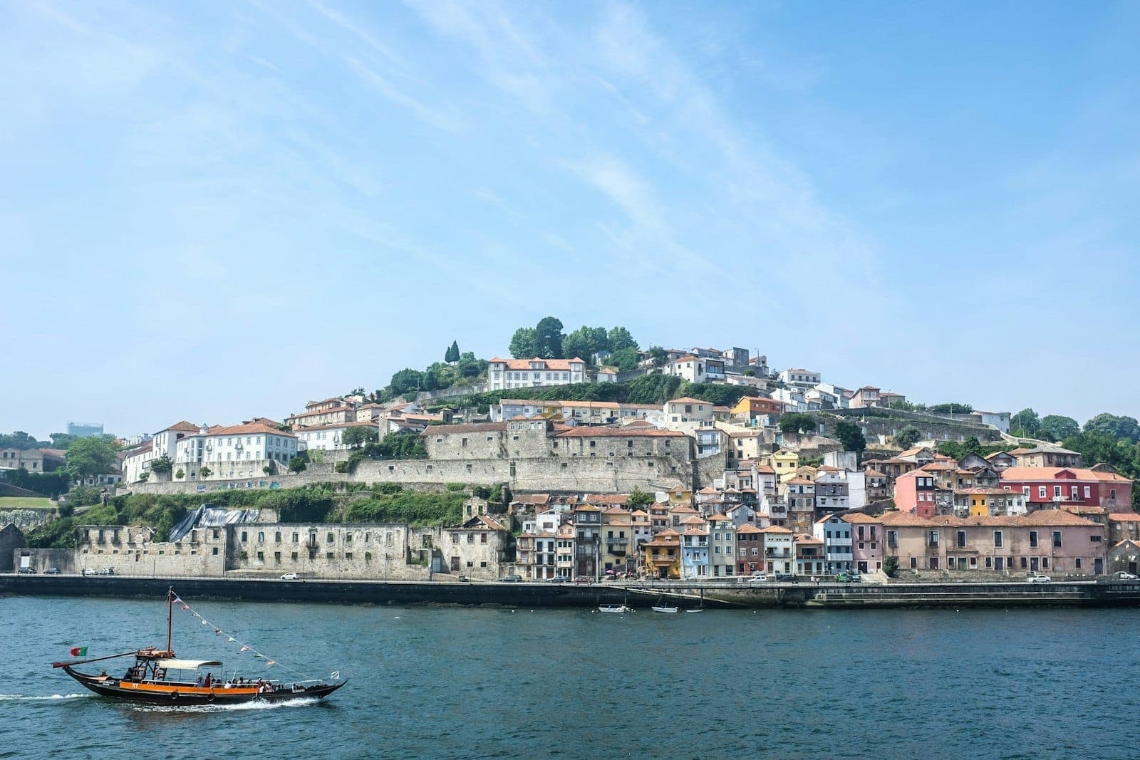 Widok na zbocze Vila Nova de Gaia, historyczne budynki i rzekę Douro z tradycyjną łodzią rabelo płynącą w słoneczny dzień.