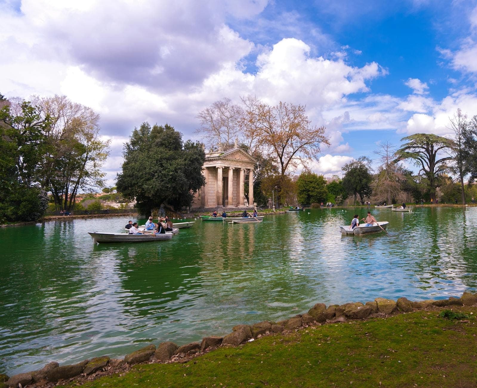 Lawa para sa pagsasagwan na may mga taong sumasakay sa bangka sa harap ng neoclassical na templo at malusog na mga puno sa Villa Borghese Gardens sa maliwanag na araw na may kaunting ulap.