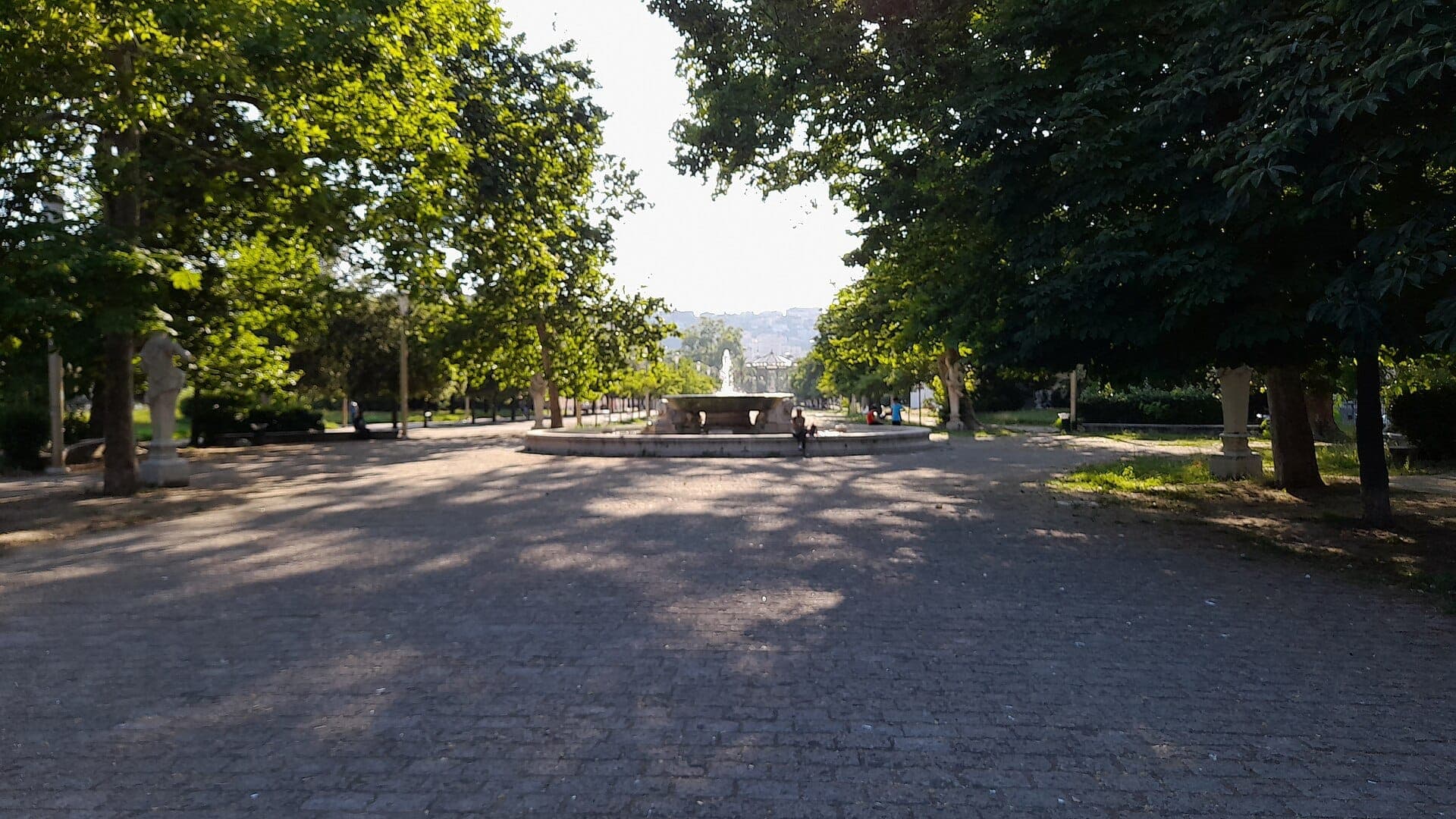 Cena ensolarada no parque com uma fonte central, alamedas arborizadas, bancos e pessoas relaxando na Villa Comunale, em Nápoles.