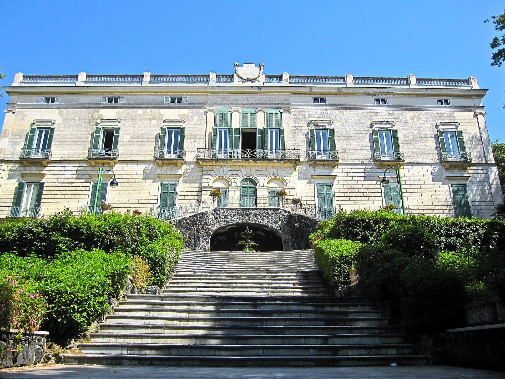 Uma ampla escadaria de pedra sobe por uma vegetação exuberante até a Villa Floridiana neoclássica com venezianas verdes e céu azul ao fundo.