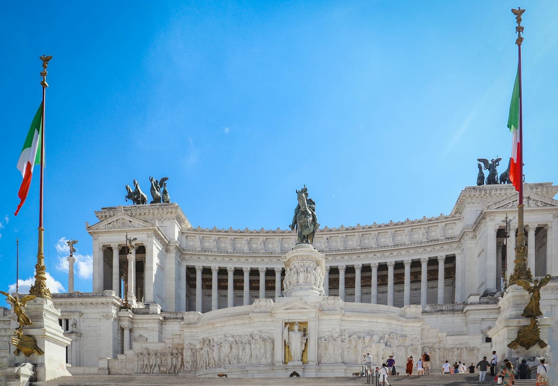 Harapang tanawin ng monumento ng Vittoriano sa Roma na may dalawang watawat ng Italya, asul na langit, puting marmol, at mga bisita sa mga hagdan.