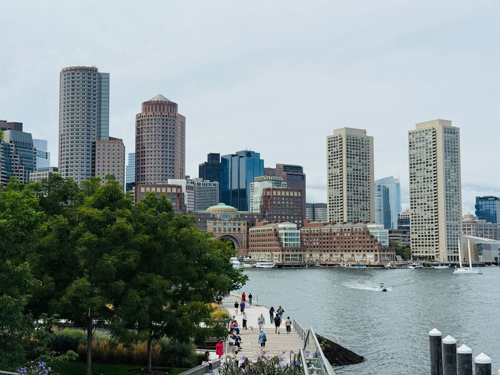 Vue de la skyline du front de mer de Boston avec des personnes se promenant sur un chemin au bord de l'eau, des arbres au premier plan et le port en arrière-plan.