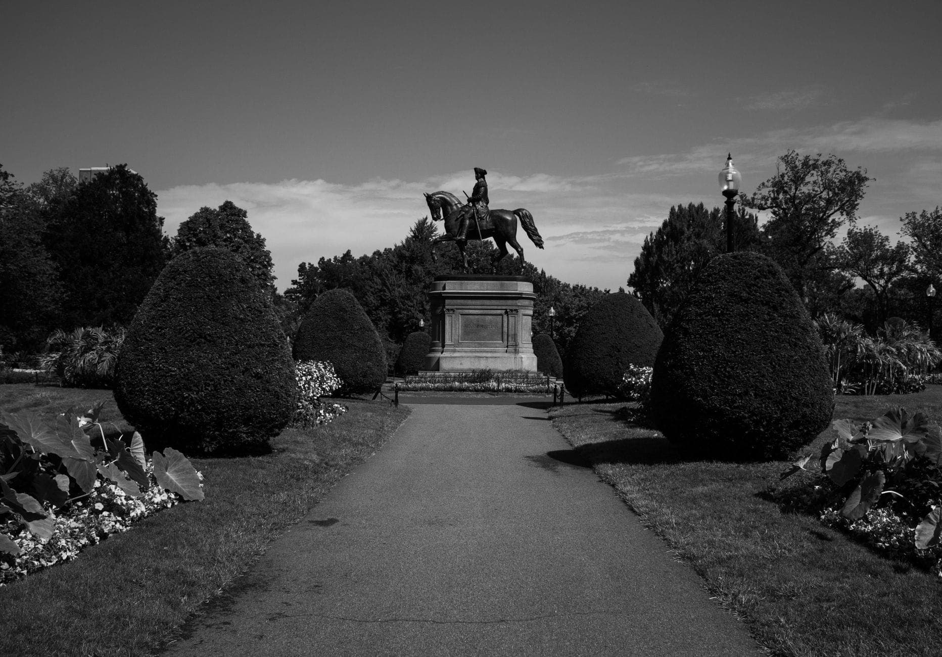Une statue d'un homme à cheval le long d'une allée bordée de jardins dans le Public Garden de Boston sous un ciel lumineux.