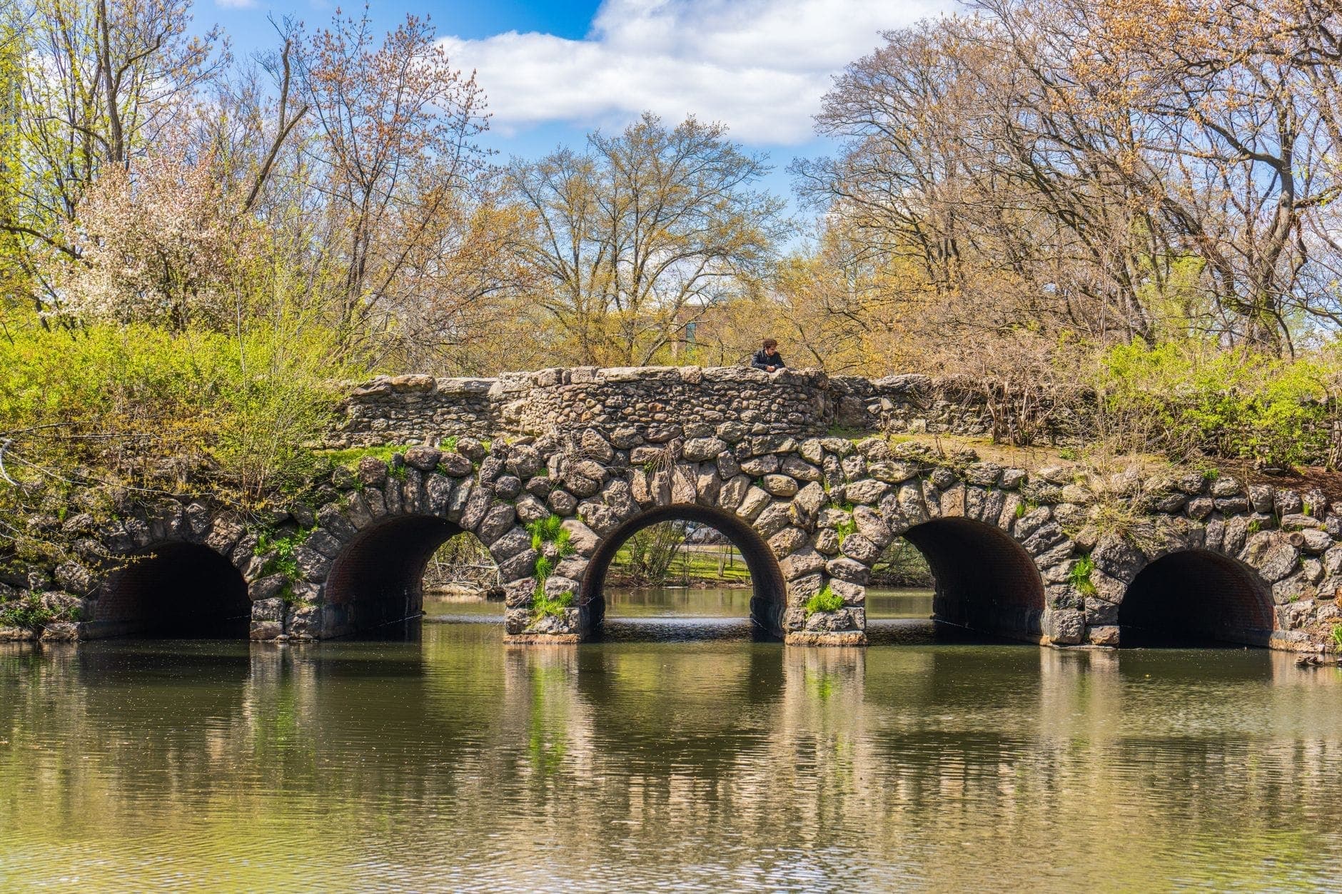 Pont en pierre à arches multiples enjambant un étang paisible dans un parc verdoyant au printemps.
