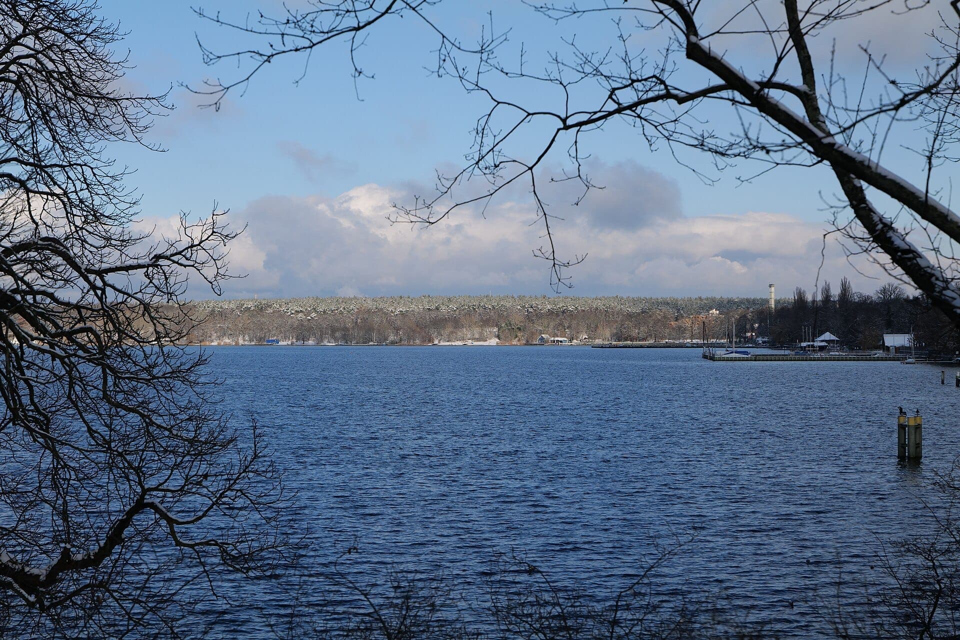 วิวทะเลสาบ Großer Wannsee ที่มีน้ำสีฟ้า กิ่งไม้ในเบื้องหน้า และอาคารบริเวณชายฝั่งฝั่งตรงข้ามภายใต้ท้องฟ้าที่มีเมฆบางส่วน