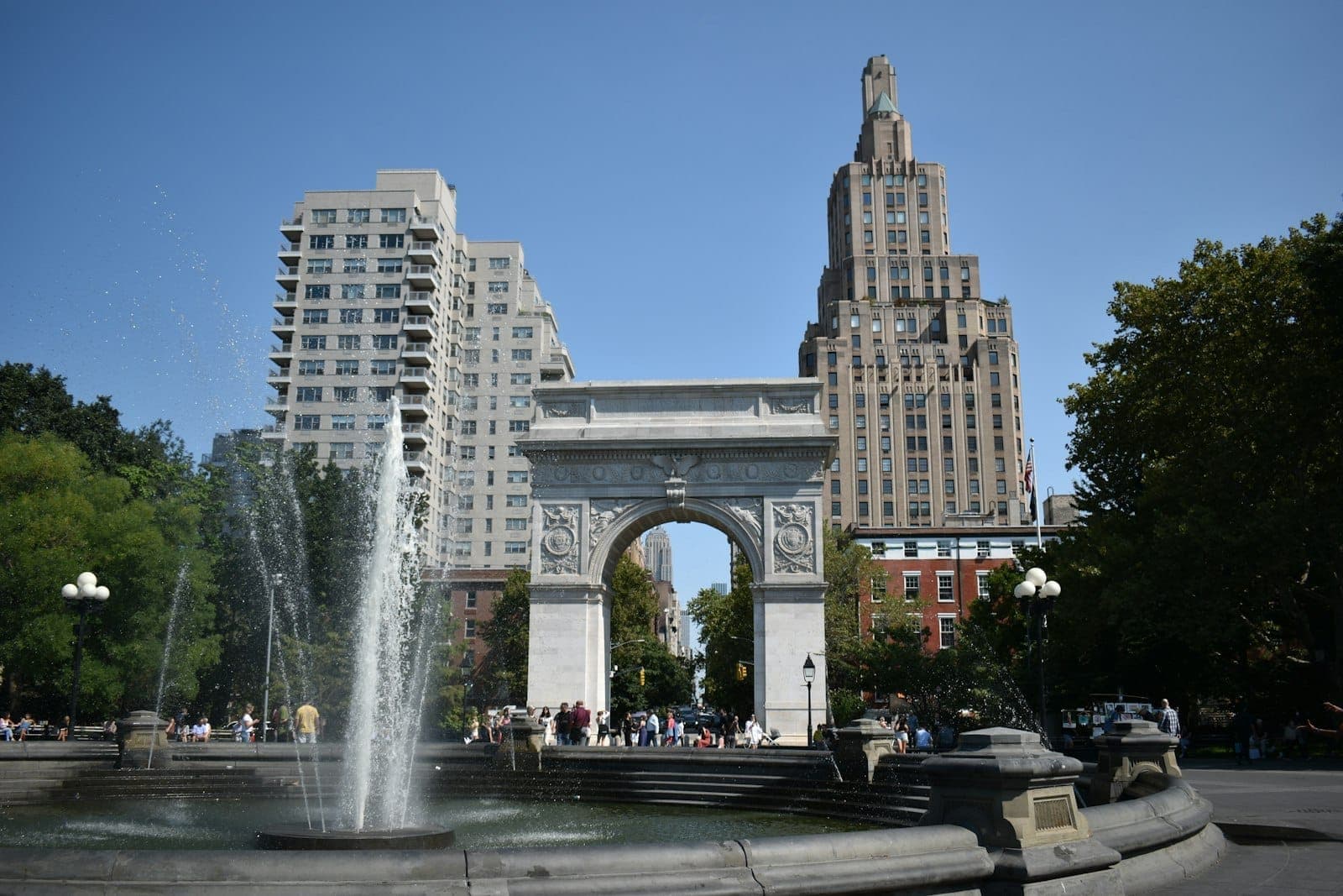 ภาพวิวซุ้มประตูหินอ่อนอันเป็นสัญลักษณ์ของ Washington Square Park กับน้ำพุใหญ่ตรงกลาง รายล้อมด้วยผู้คน ต้นไม้ และตึกเก่าแก่ Greenwich Village ท่ามกลางท้องฟ้าใส