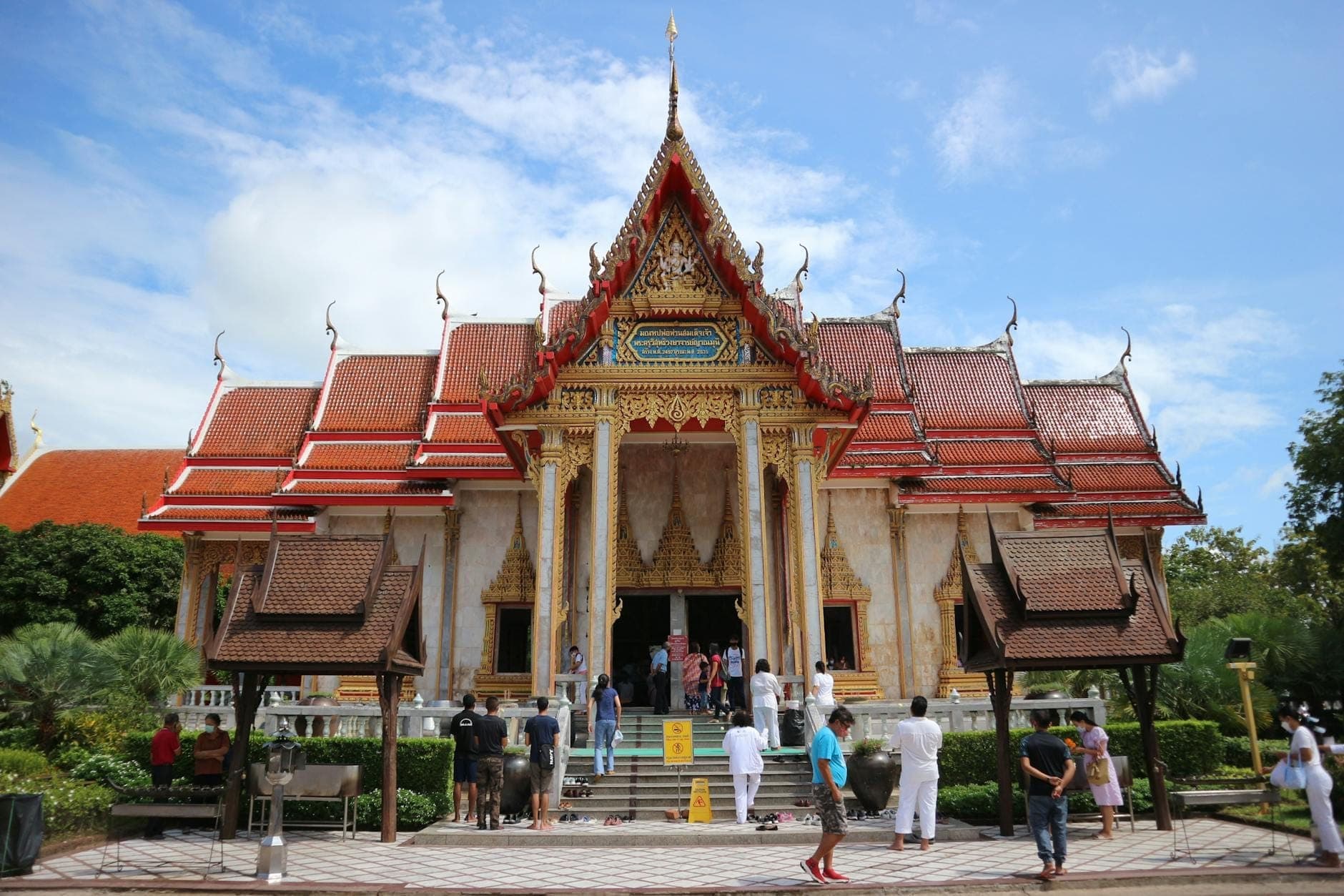 Front view of Wat Chalong temple in Phuket with ornate red and gold roof, intricate carvings, and visitors entering under a blue sky.