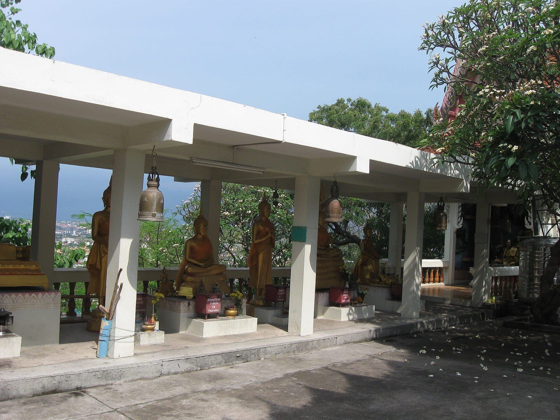 Золотые статуи Будды под белым павильоном в Ват Као Пра Бат, окружённые зеленью на фоне залива Паттайя.