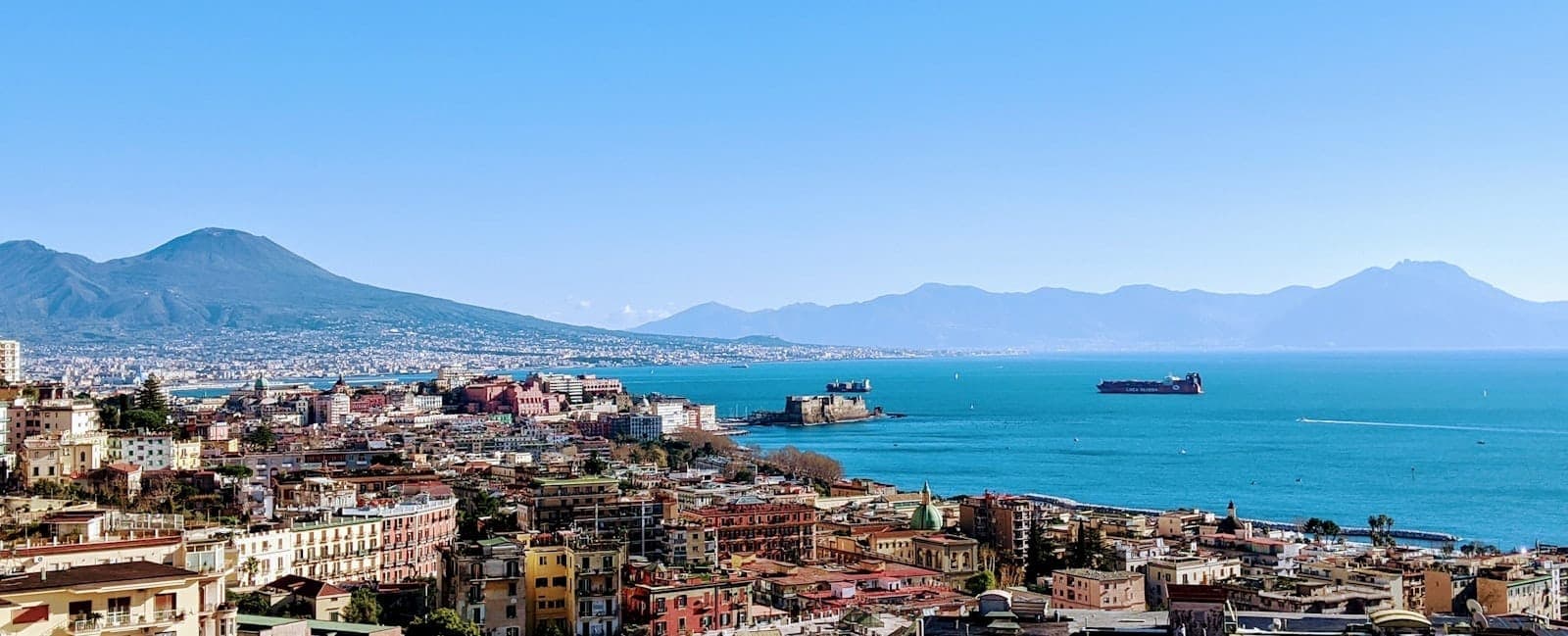 Vista panorâmica da orla de Nápoles com o Lungomare, a Baía de Nápoles, o Castel dell'Ovo e o Monte Vesúvio ao fundo sob um céu azul limpo.