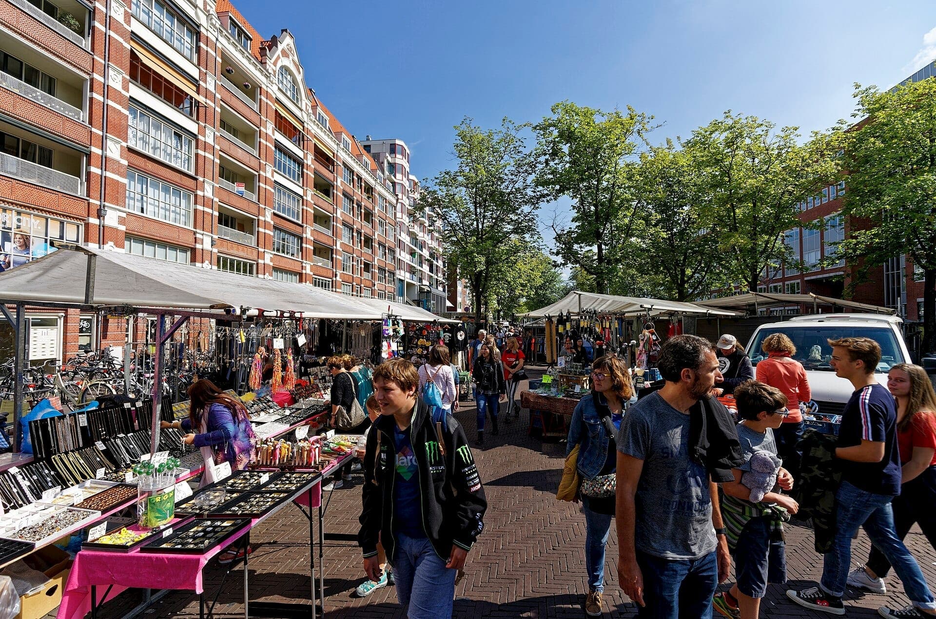 Reihen von Marktständen und Einkäufern am Waterlooplein Market in Amsterdam, mit Backsteinwohngebäuden und belaubten Bäumen unter einem strahlend blauen Himmel.