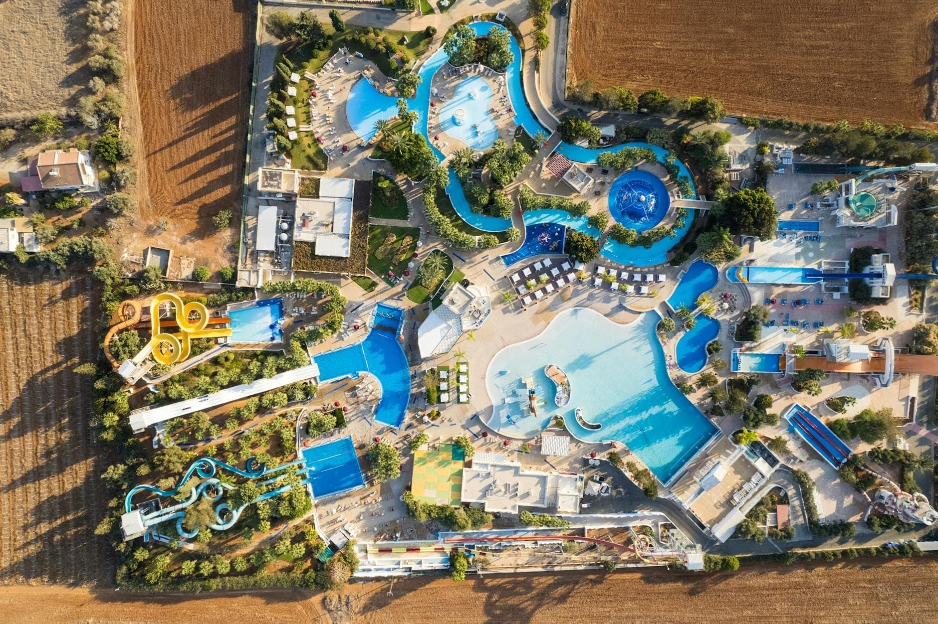 Vue aérienne du Waterpark Faliraki avec ses toboggans colorés, ses piscines sinueuses et ses espaces de bronzage entourés d'arbres et de champs à Rhodes, en Grèce.
