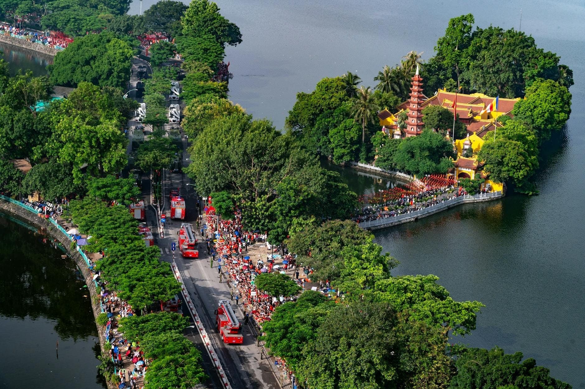 Widok z lotu ptaka na West Lake w Hanoi z Pagodą Tran Quoc otoczoną bujnymi drzewami, tłumami i wozami strażackimi wzdłuż drogi nad jeziorem.