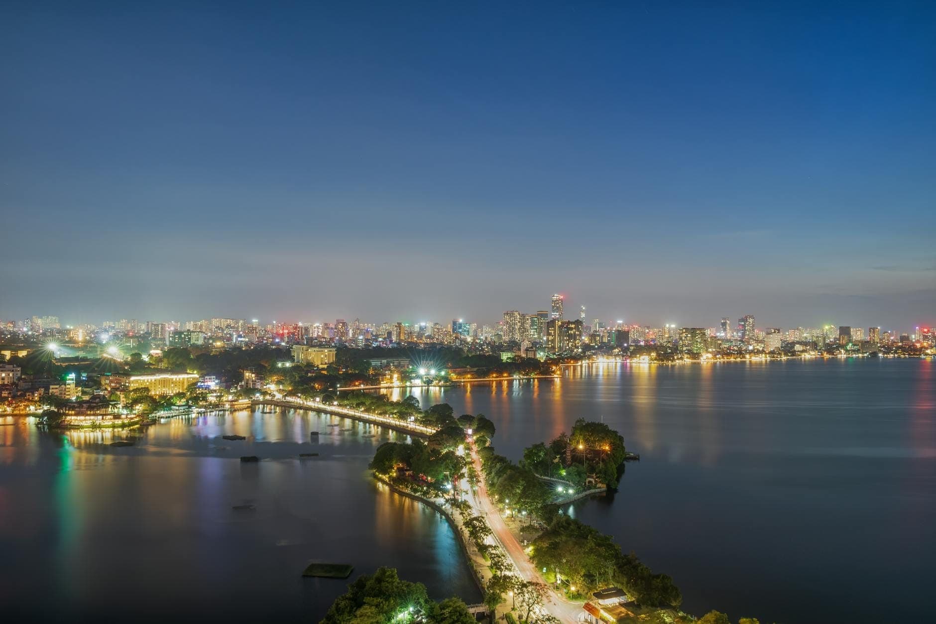 Zapierający dech w piersiach widok z lotu ptaka na Zachodnie Jezioro w Hanoi o zachodzie słońca – kręta aleja obsadzona drzewami, światła miasta i spokojna woda odbijająca barwy wieczornego nieba.