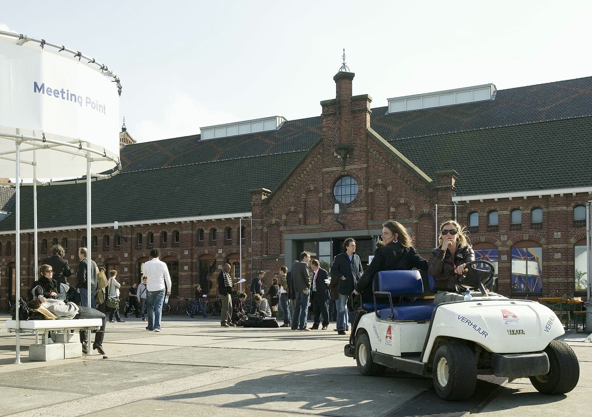 Menschengruppen versammeln sich vor dem historischen Backsteingebäude des Westergasfabriek in Amsterdam unter einem klaren Himmel, während ein Schild mit der Aufschrift 'Meeting Point' sichtbar ist.