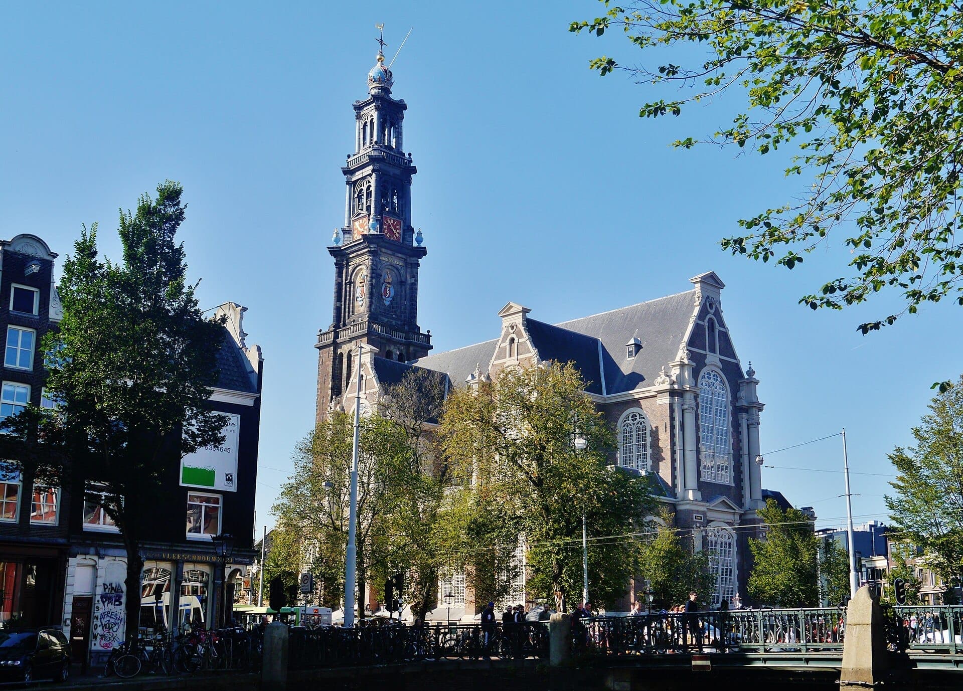 Die Westerkerk-Kirche in Amsterdam ragt neben dem Prinsengracht-Kanal empor, umgeben von Bäumen und klassischen niederländischen Gebäuden an einem sonnigen Tag.