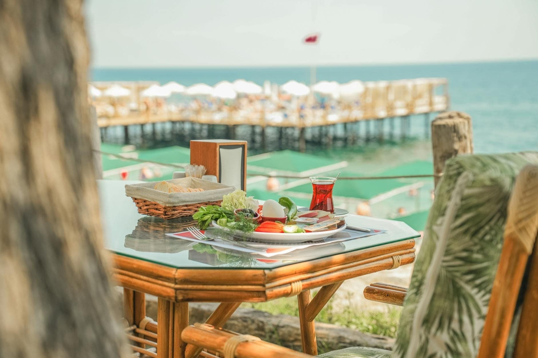Frühstückstisch mit türkischem Tee, Gemüse und Brot im Freien mit Meerblick in Antalya, mit einem Steg und Sonnenschirmen im Blick.