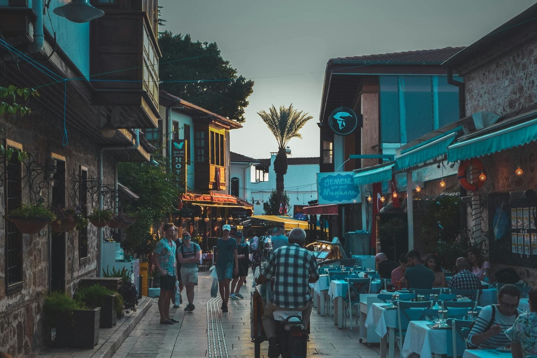 Belebte, von Restaurants gesäumte Straße in Antalya in der Abenddämmerung mit Einheimischen und Besuchern, die draußen speisen und an traditionellen Gebäuden vorbeigehen.