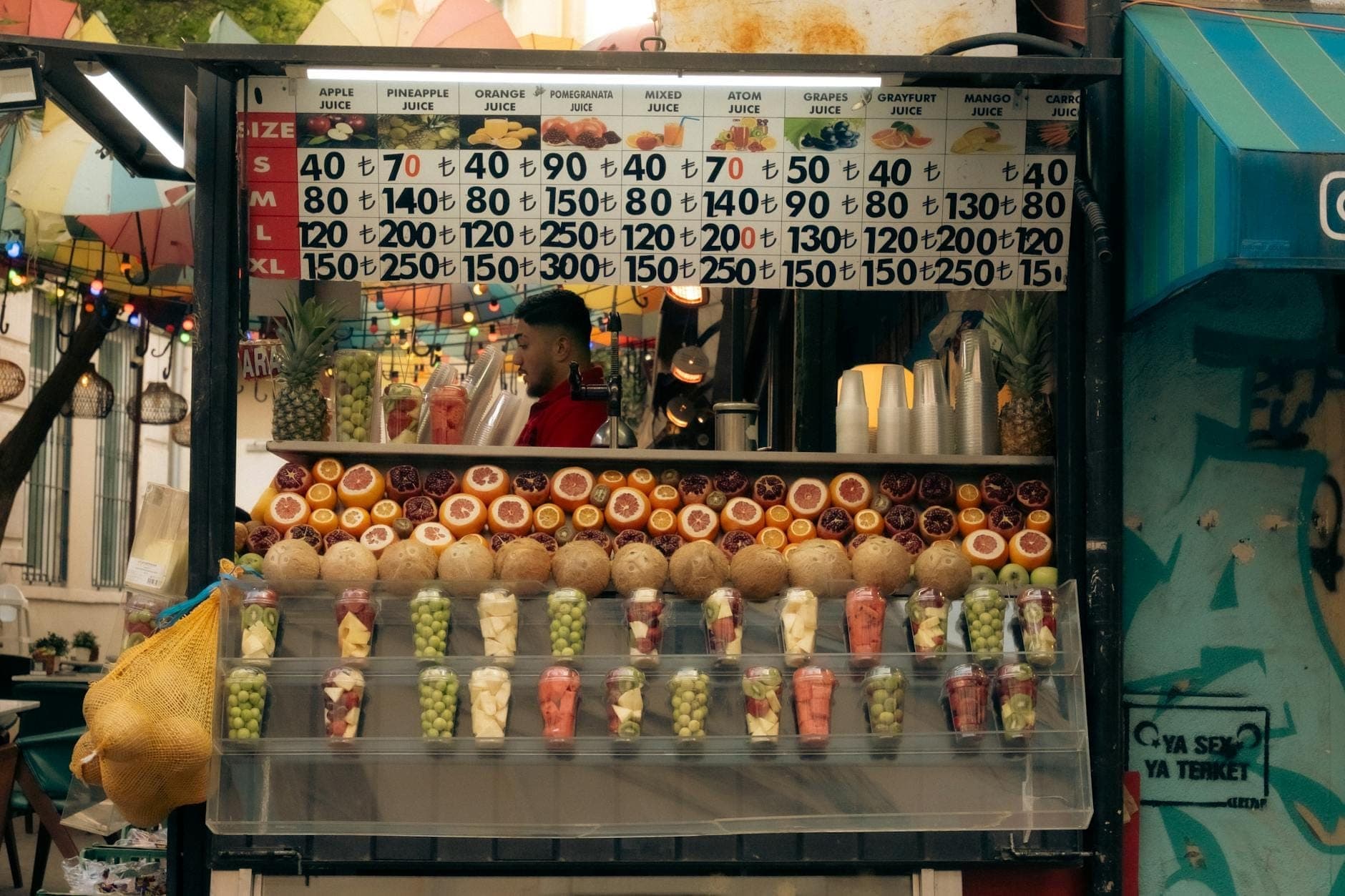 Ein bunter Saftstand in einer Straßenumgebung mit frischen Orangen, Granatäpfeln, Ananas und Bechern mit gemischten Früchten, der eine Vielzahl von Getränken in Antalya anbietet.