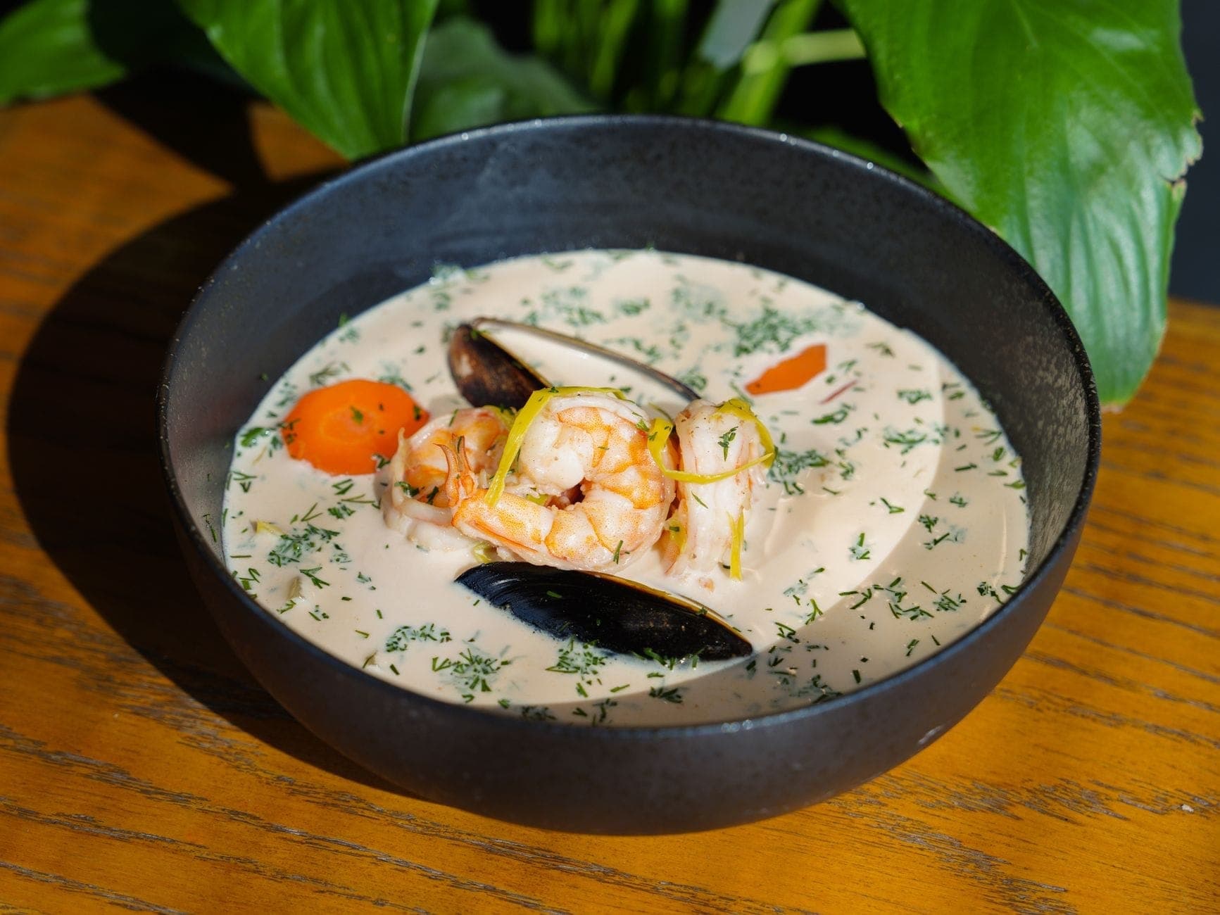 Un bol de chowder crémeux aux fruits de mer garni de crevettes, moules, herbes et légumes sur une table en bois.